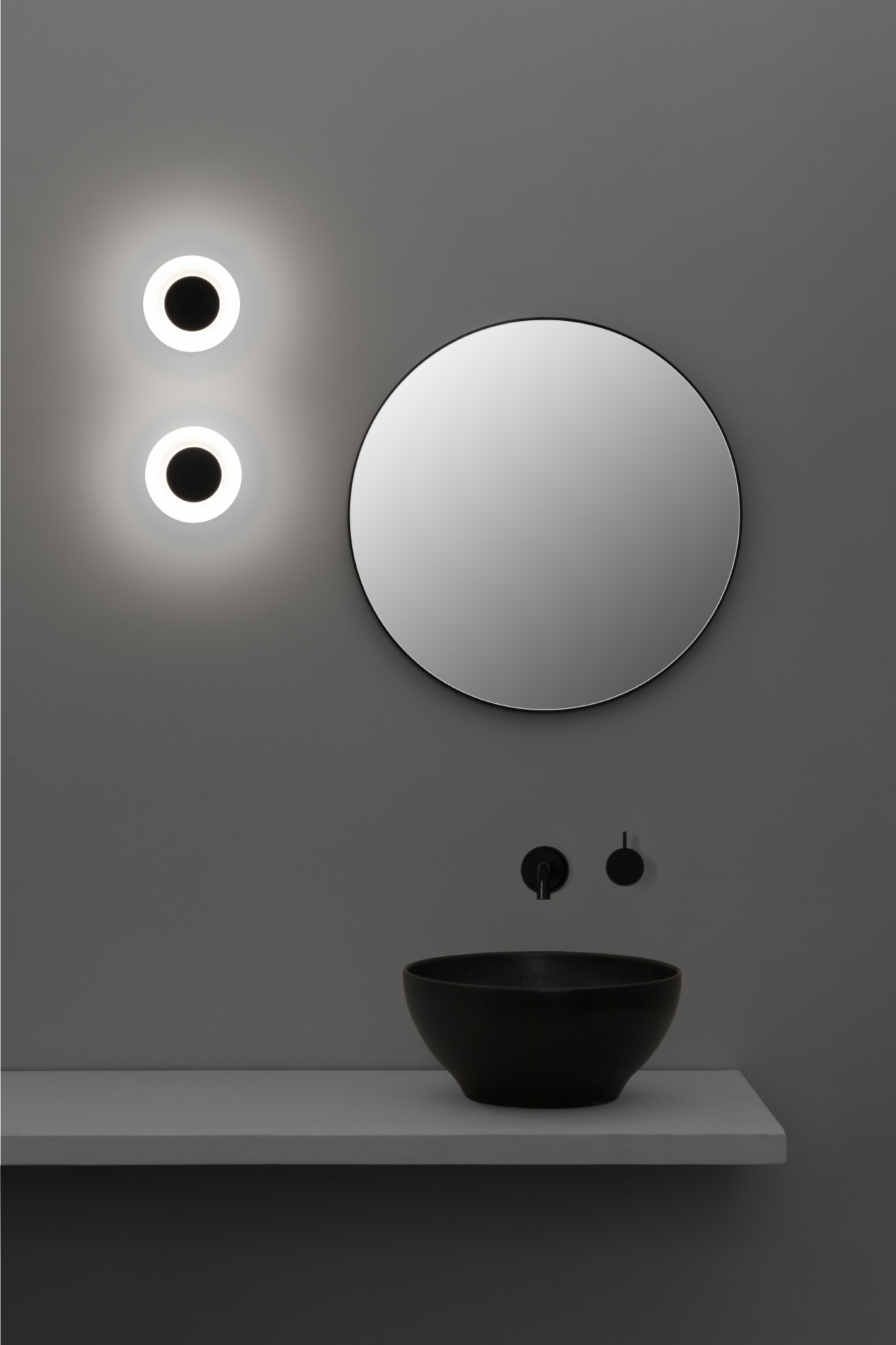 Interni del bagno. Specchio rotondo, lavabo nero, rubinetto e due luci circolari. Parete grigia e mensola bianca.
