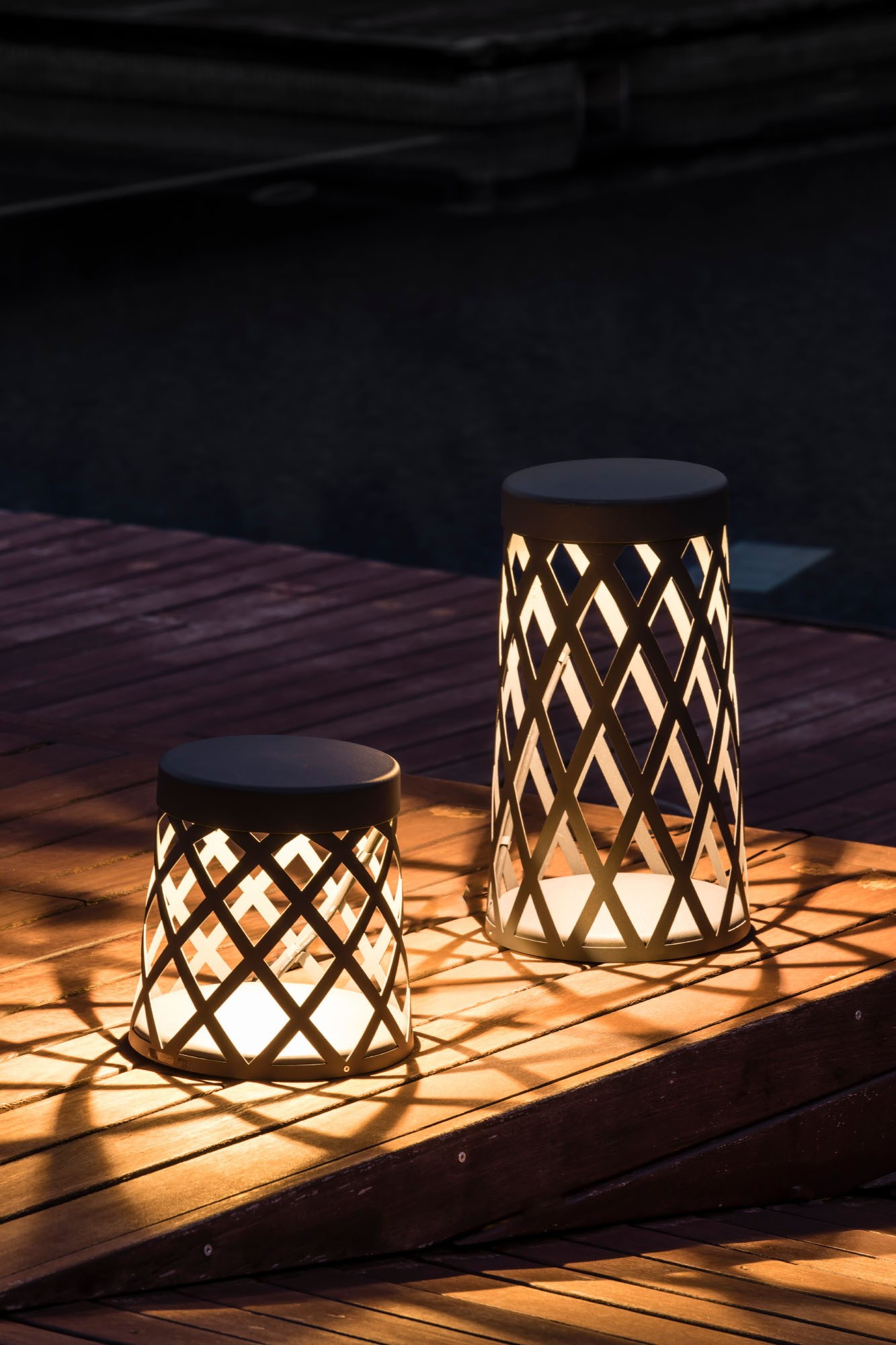 Due lampade da esterno su una terrazza in legno. Le lampade hanno un design a griglia ed emettono un bagliore caldo. Sfondo scuro.