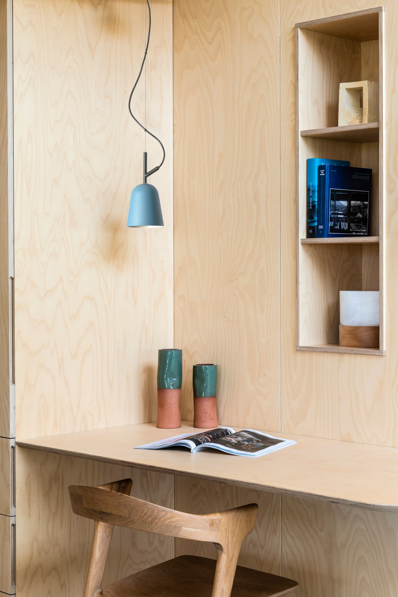 Una scrivania in legno con sedia, libri e vasi. Una lampada blu è appesa sopra. Lo sfondo è composto da pannelli di legno.