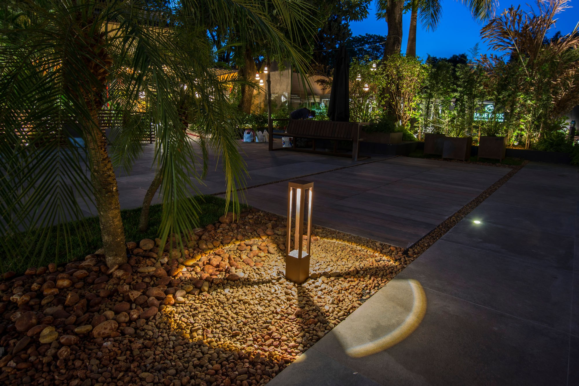 Scena notturna con palme, un sentiero illuminato e una panchina. Un moderno apparecchio di illuminazione illumina ghiaia e pietre.