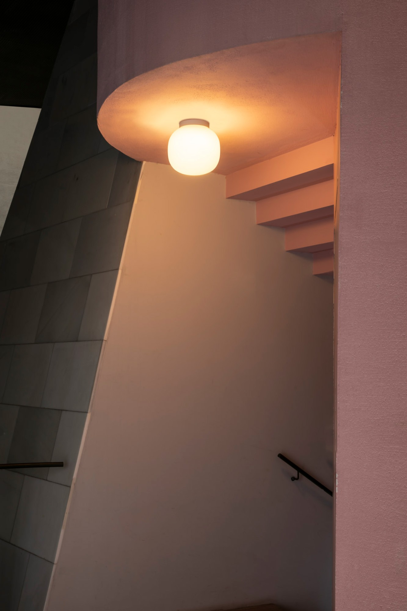 Scatto interno di scale con una luce rotonda luminosa. Pareti e gradini rosa, corrimano scuro. Parete di piastrelle grigie a sinistra.
