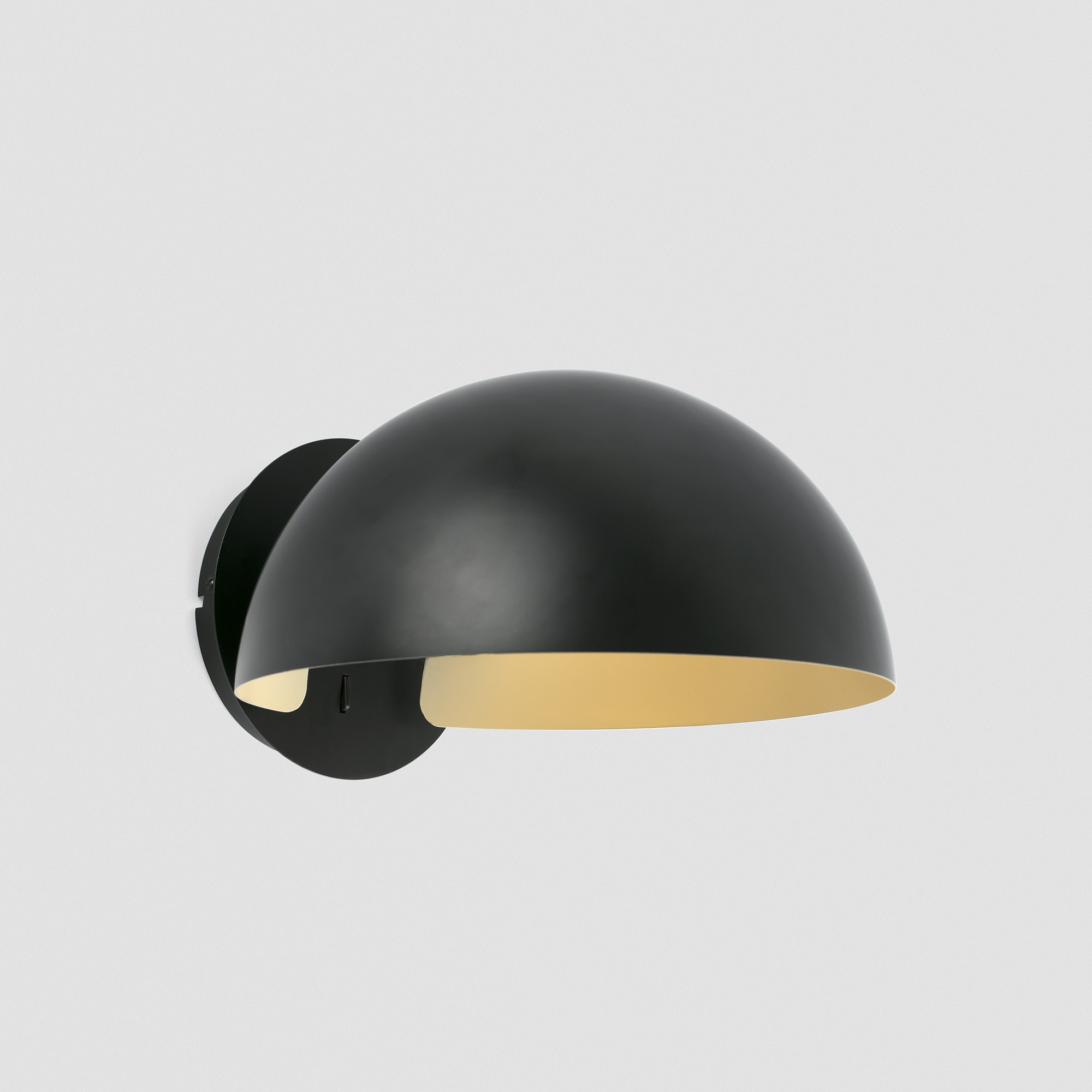 Una lampada da parete nera a forma di cupola con interno dorato. La lampada è montata su un muro bianco.