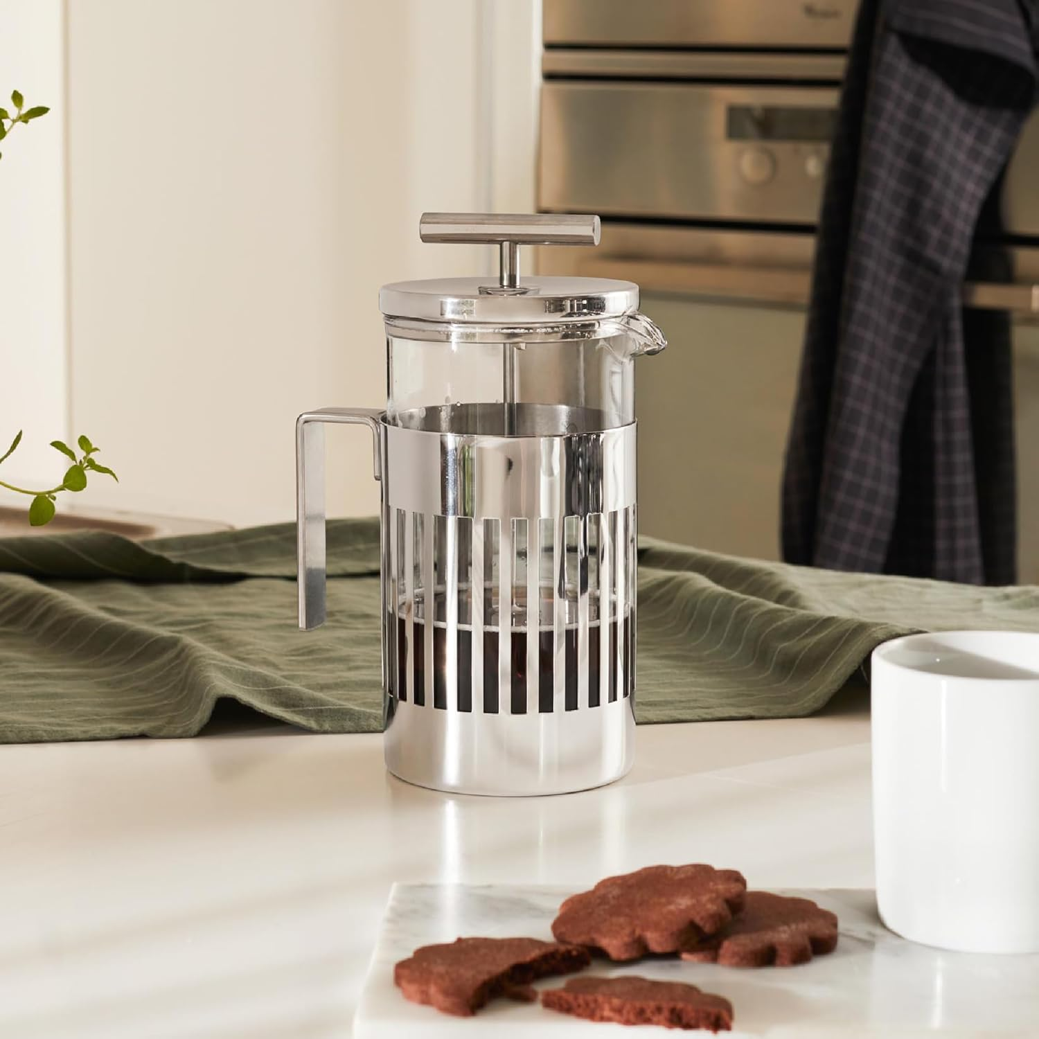 Caffettiera francese, biscotti e tazza su una superficie bianca. Panno verde e forno sullo sfondo. La pressa ha caffè dentro.