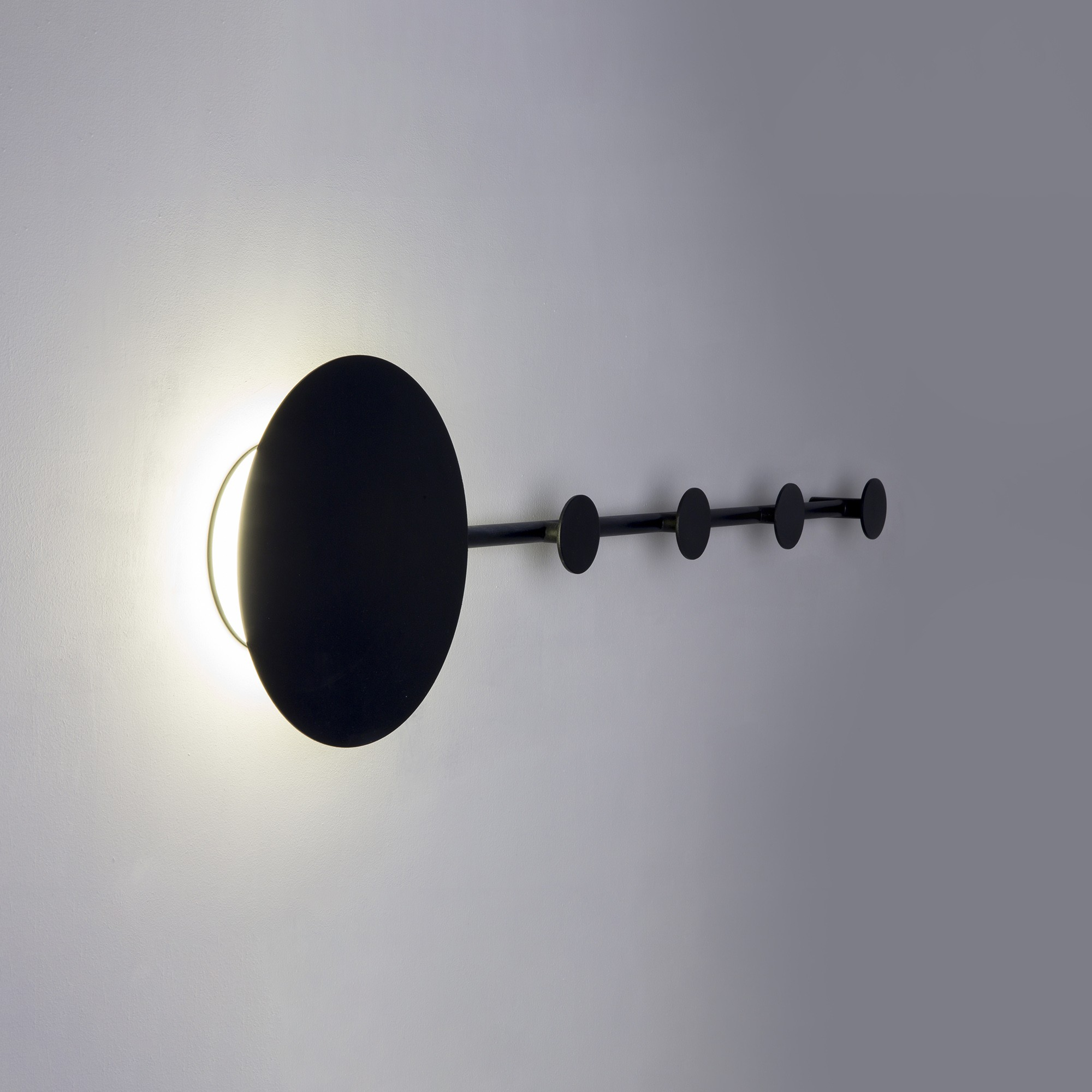 Una moderna lampada da parete nera con un design circolare e cinque piccoli ganci rotondi, contro un muro grigio.
