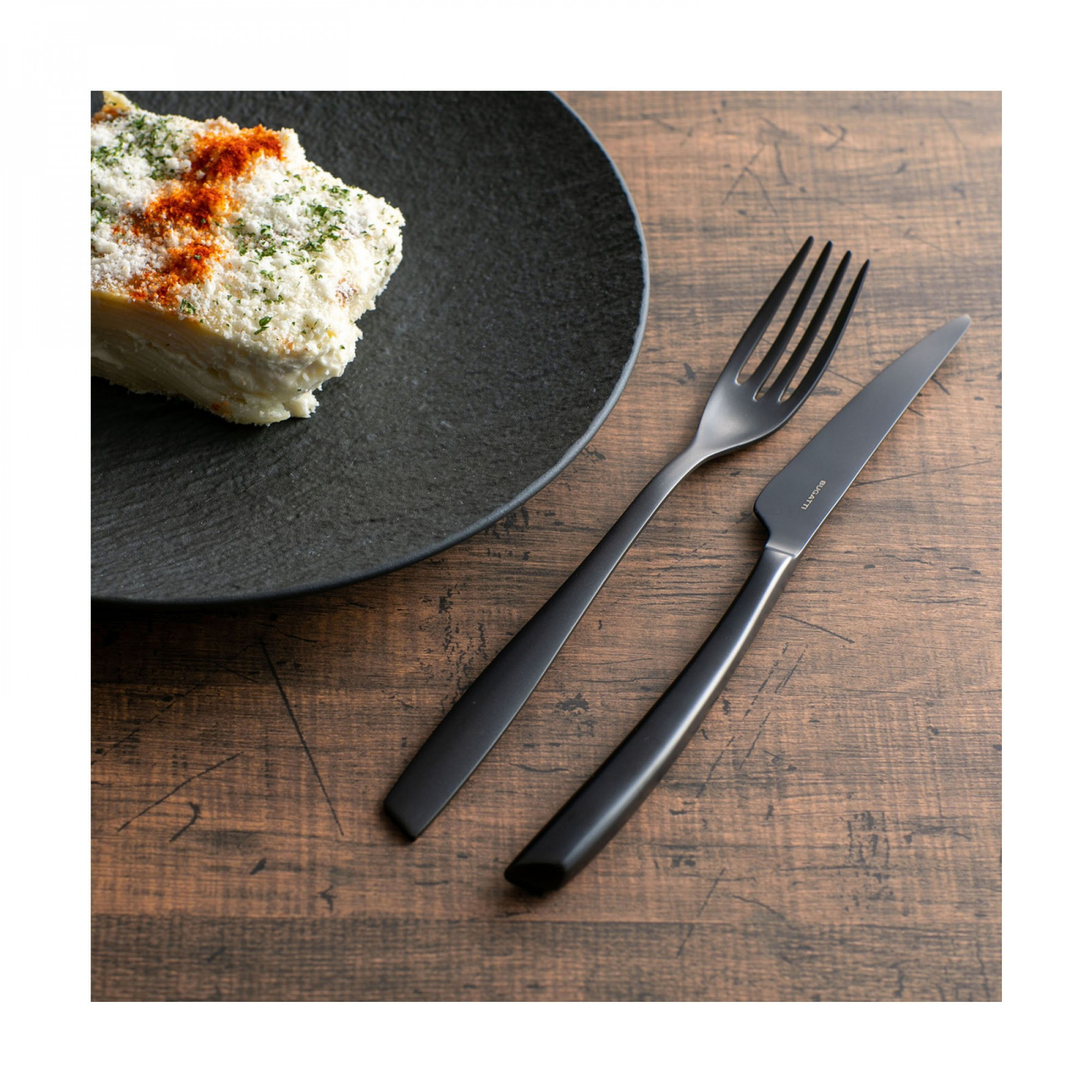 Un piatto nero con un pezzo di cibo a strati, una forchetta e un coltello su un tavolo di legno.