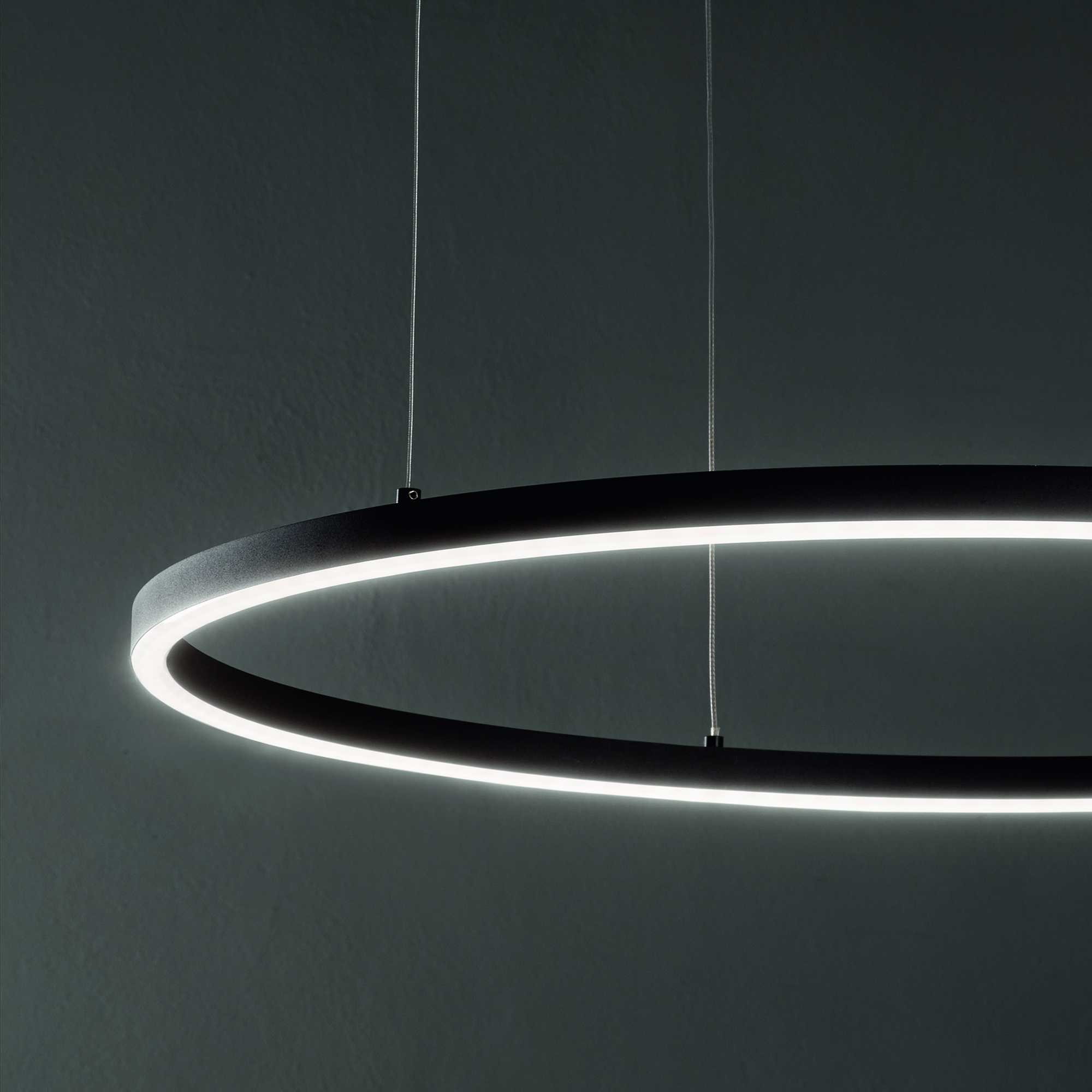 Un moderno apparecchio di illuminazione nero a forma di anello è appeso a una parete grigio scuro. La luce emette una brillante luce bianca.