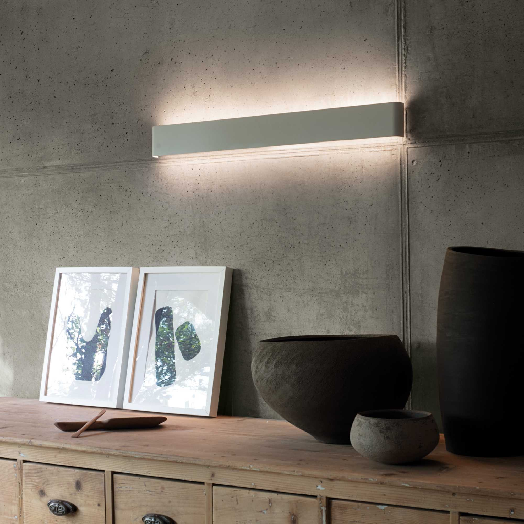 Apparecchio di illuminazione bianco rettangolare su muro di cemento. Due quadri e vasi su superficie in legno.