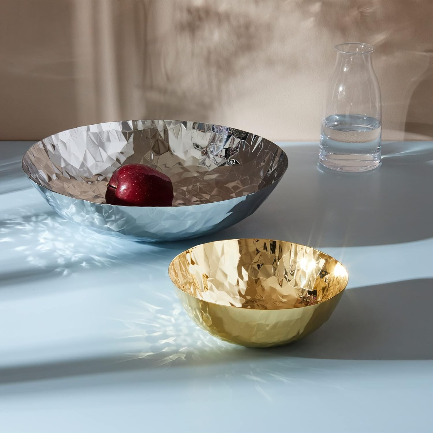 Una ciotola d'argento con una mela rossa, una ciotola d'oro e un vaso di vetro con acqua su una superficie blu, sfondo beige.