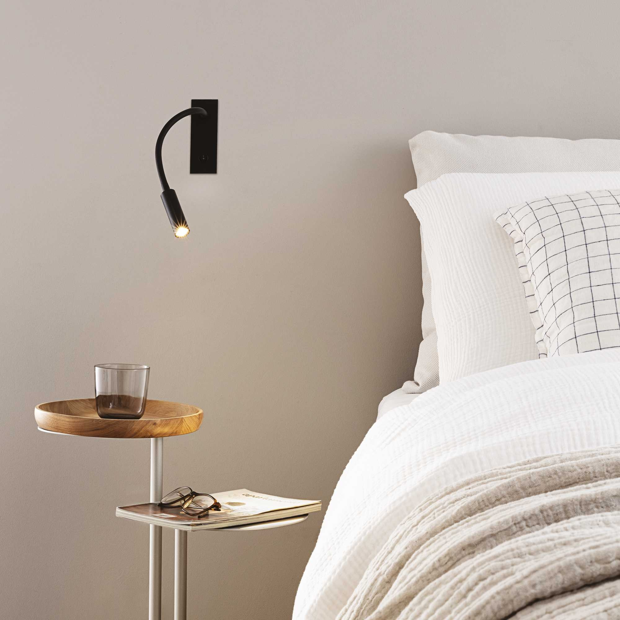 Una lampada da comodino nera illumina un tavolino. Un bicchiere, occhiali e una rivista sono sul tavolo. Un letto sullo sfondo.