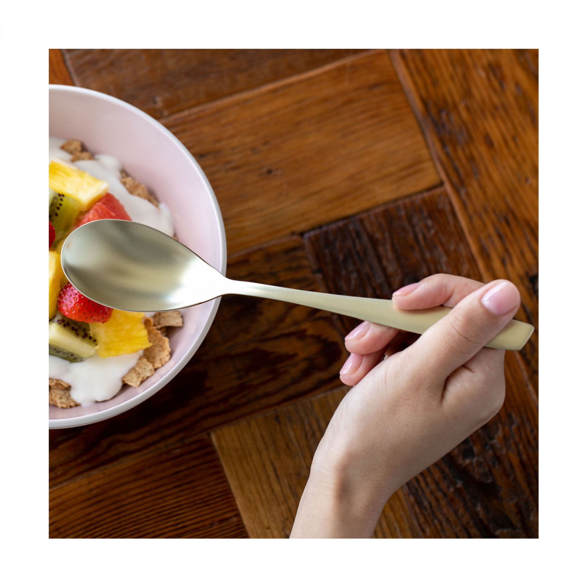Una mano tiene un cucchiaio d'oro sopra una ciotola di cereali con frutta su un tavolo di legno.