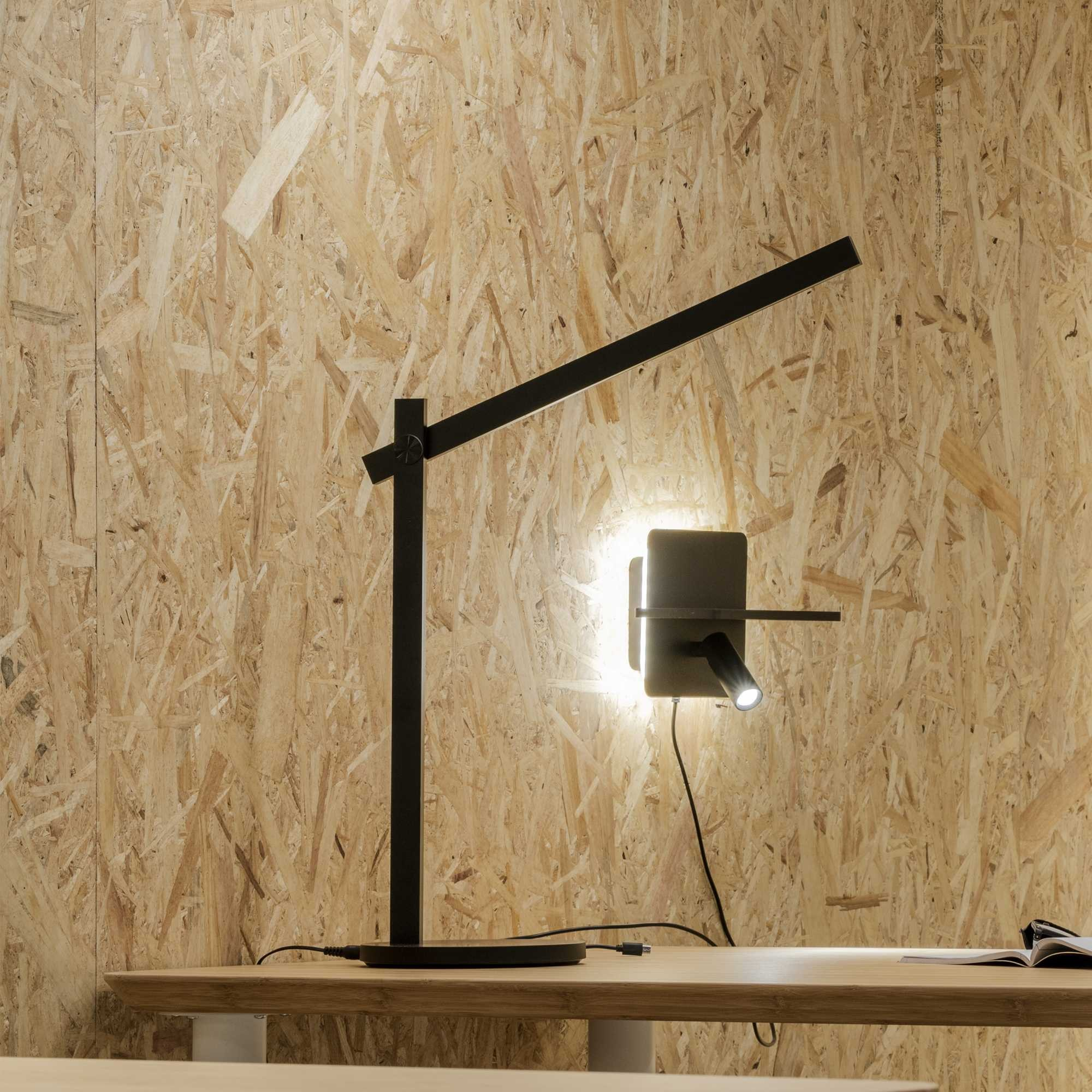 Una lampada da scrivania nera e moderna è su una scrivania di legno. Il muro è fatto di trucioli di legno.
