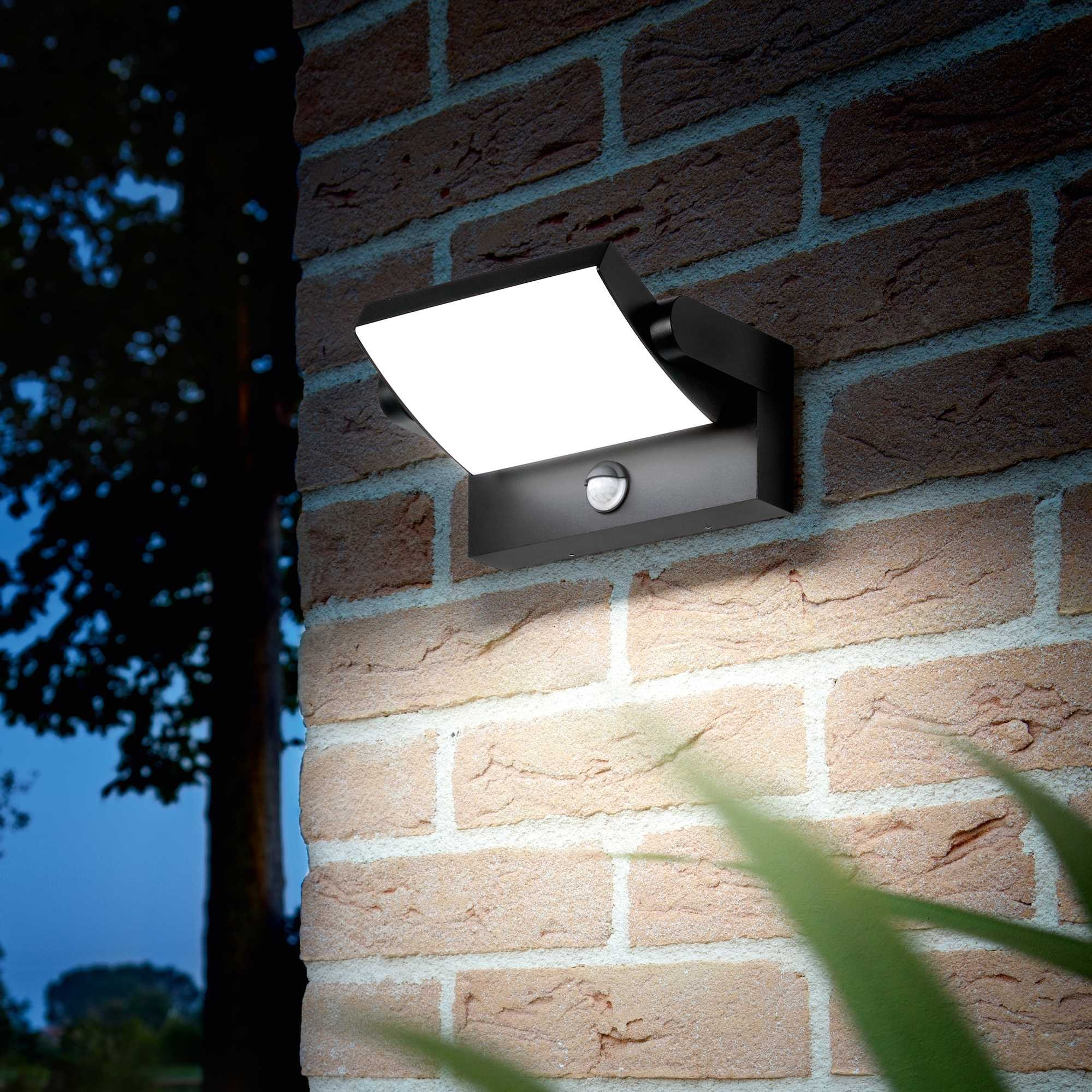 Una lampada nera da esterno su un muro di mattoni. La lampada è accesa, che illumina il muro. Scena notturna.