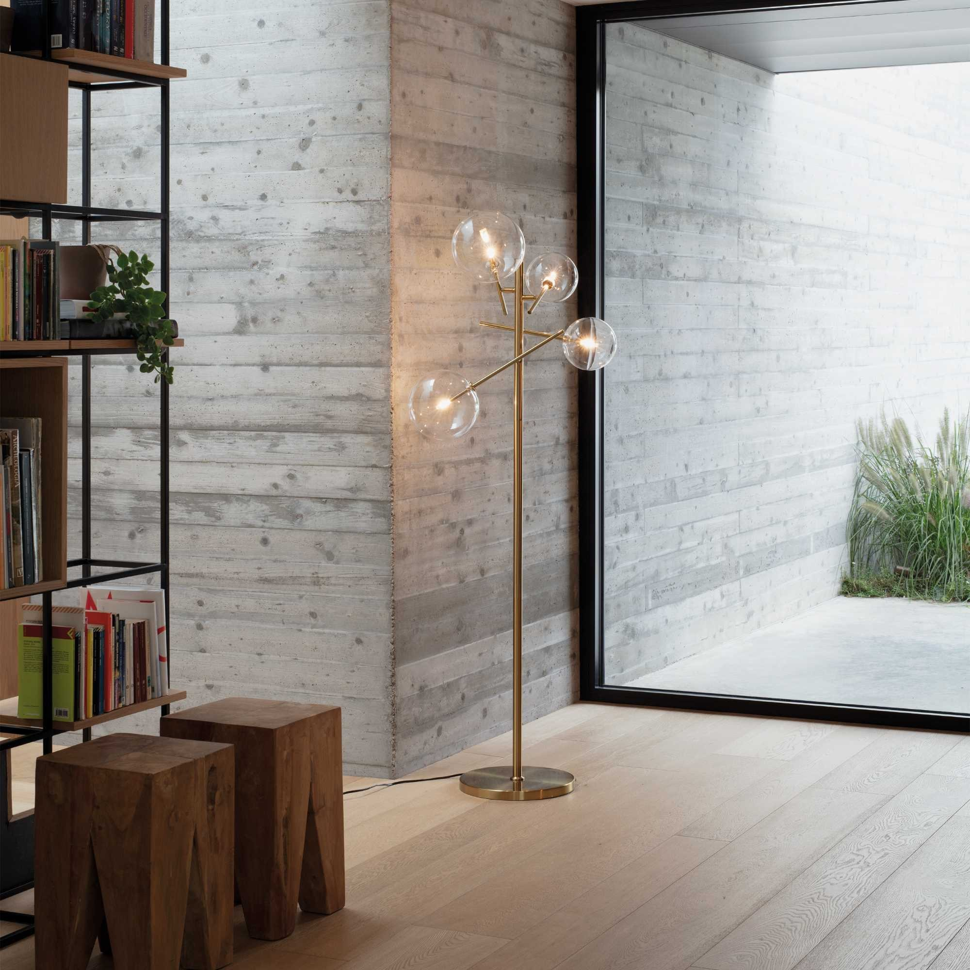 Interni moderni con lampada da terra in ottone con globi, sgabelli in legno e libreria. Parete in cemento e finestra.