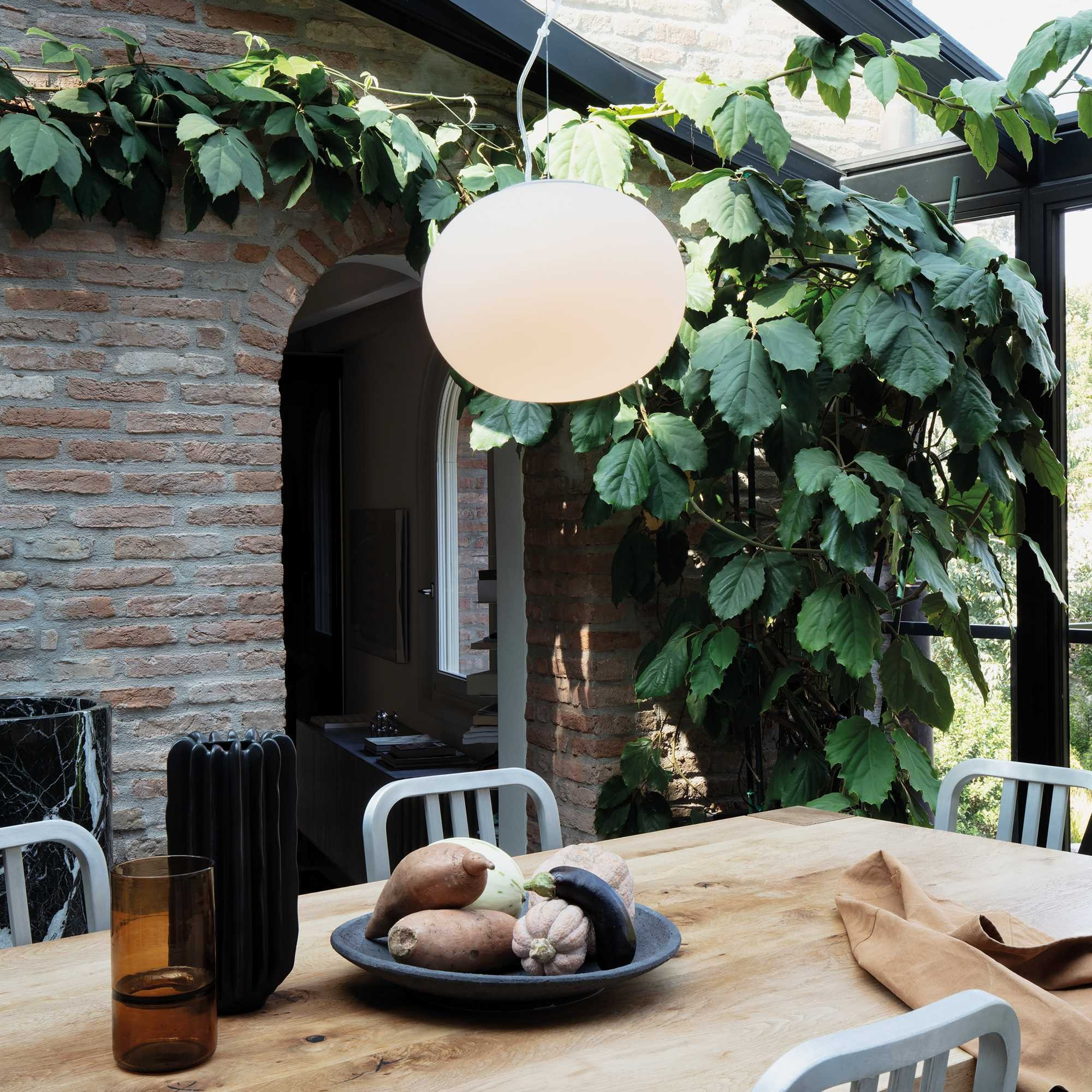 Sala da pranzo con tavolo di legno, verdure, una grande lampada a sospensione e piante rampicanti. Muri in mattoni e finestre.