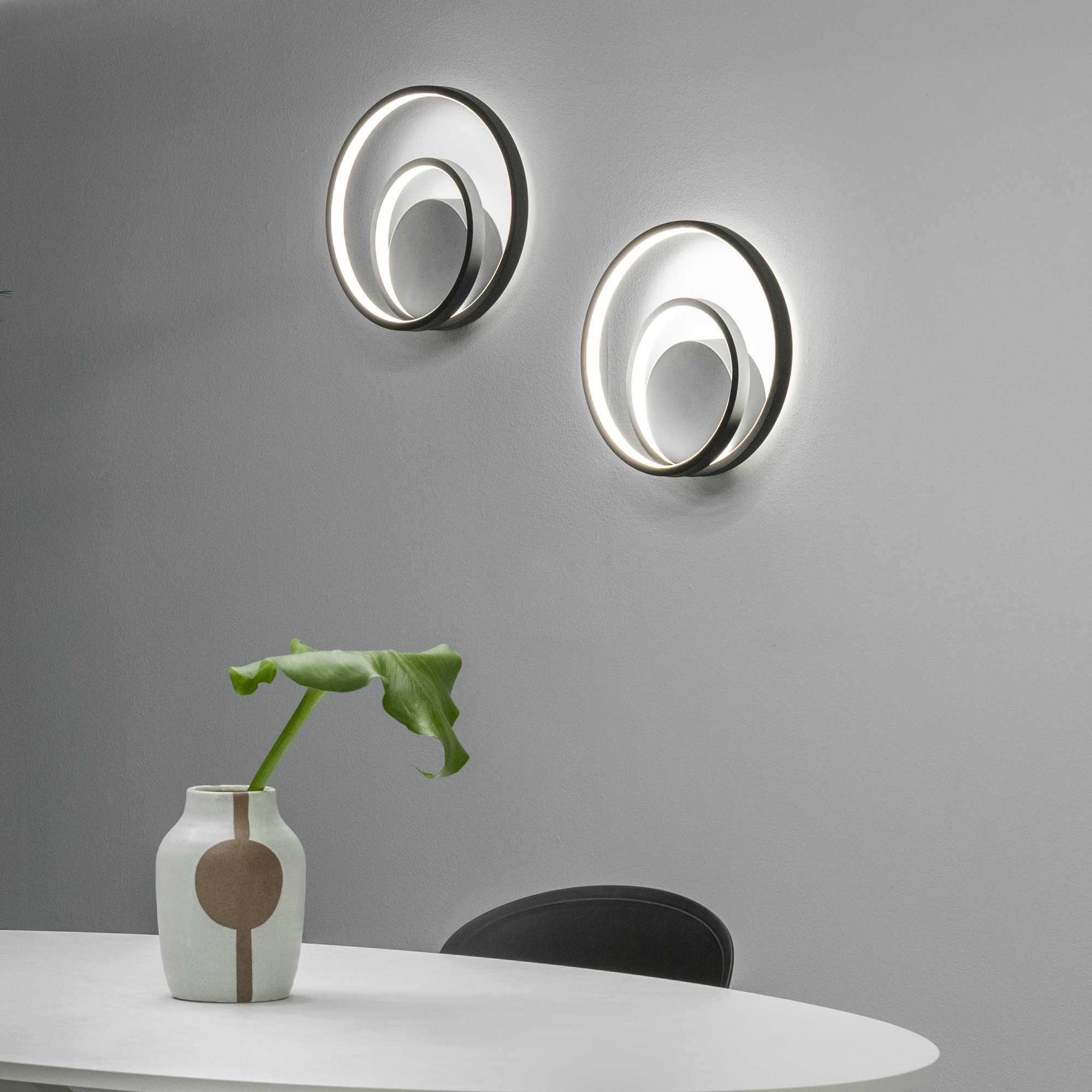 Due lampade da parete circolari illuminate. Un vaso con una pianta è su un tavolo bianco. Una sedia scura è dietro il tavolo.