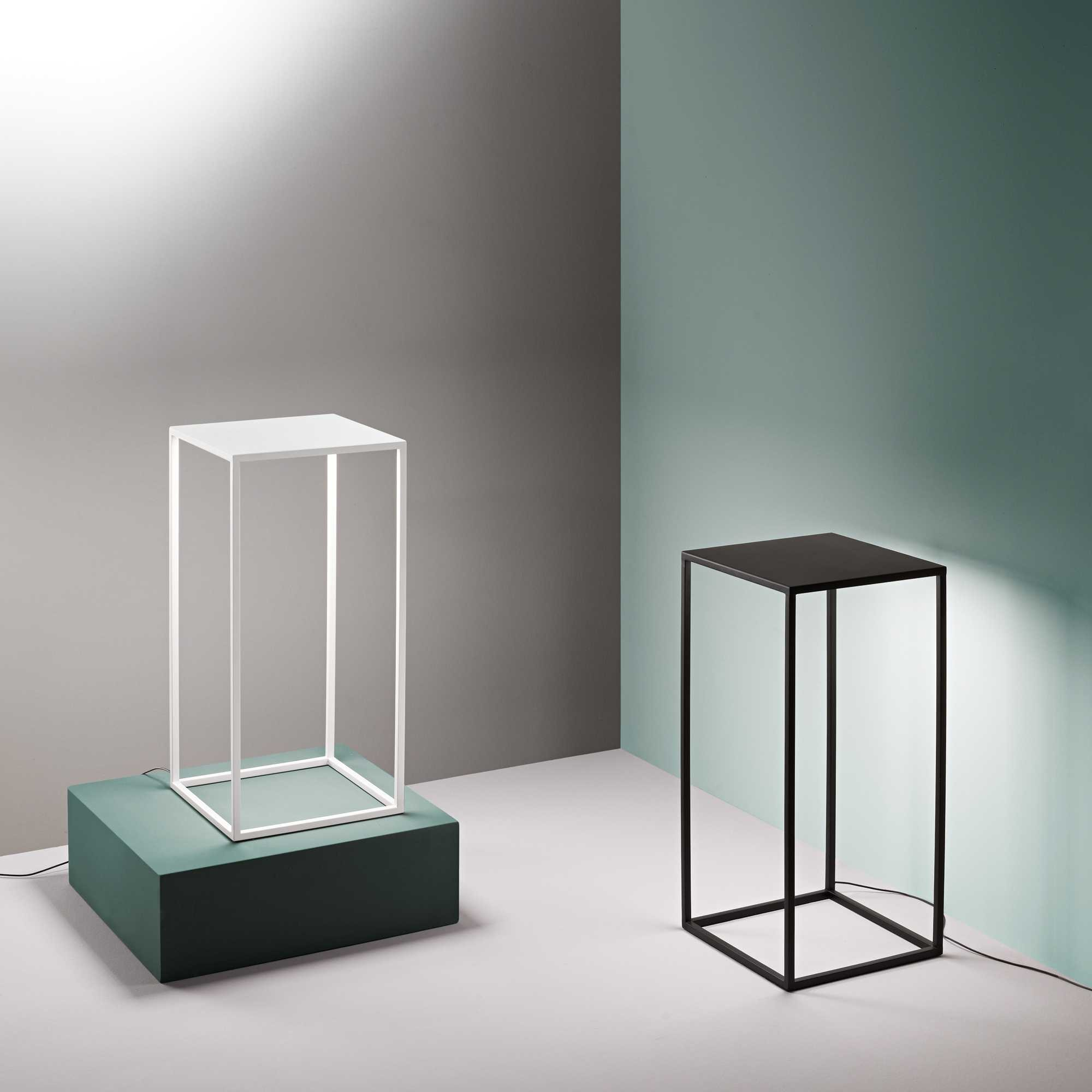 Due lampade a forma di cubo, una bianca, una nera, su sfondo grigio e verde acqua. La bianca poggia su un blocco verde.