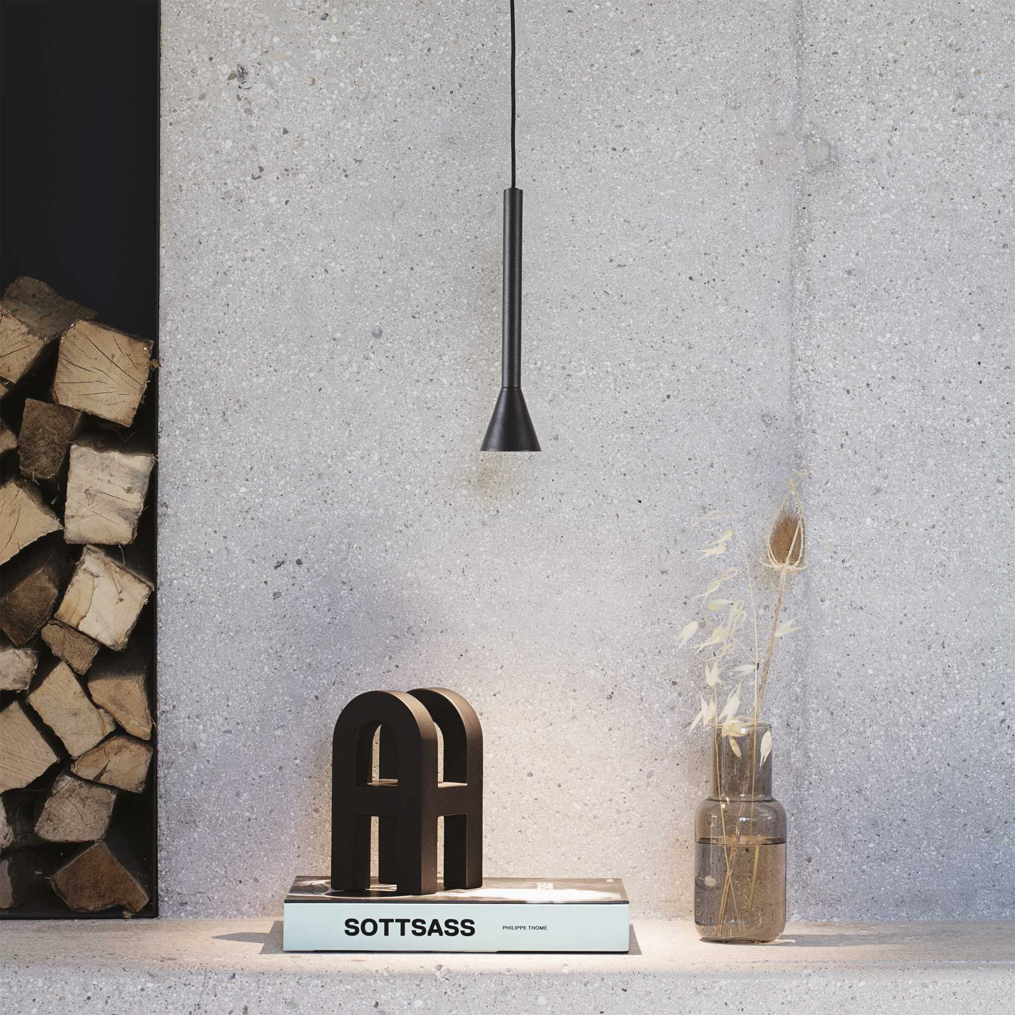 Scena interna moderna con lampada a sospensione, oggetti decorativi e una pila di legna contro un muro di cemento.