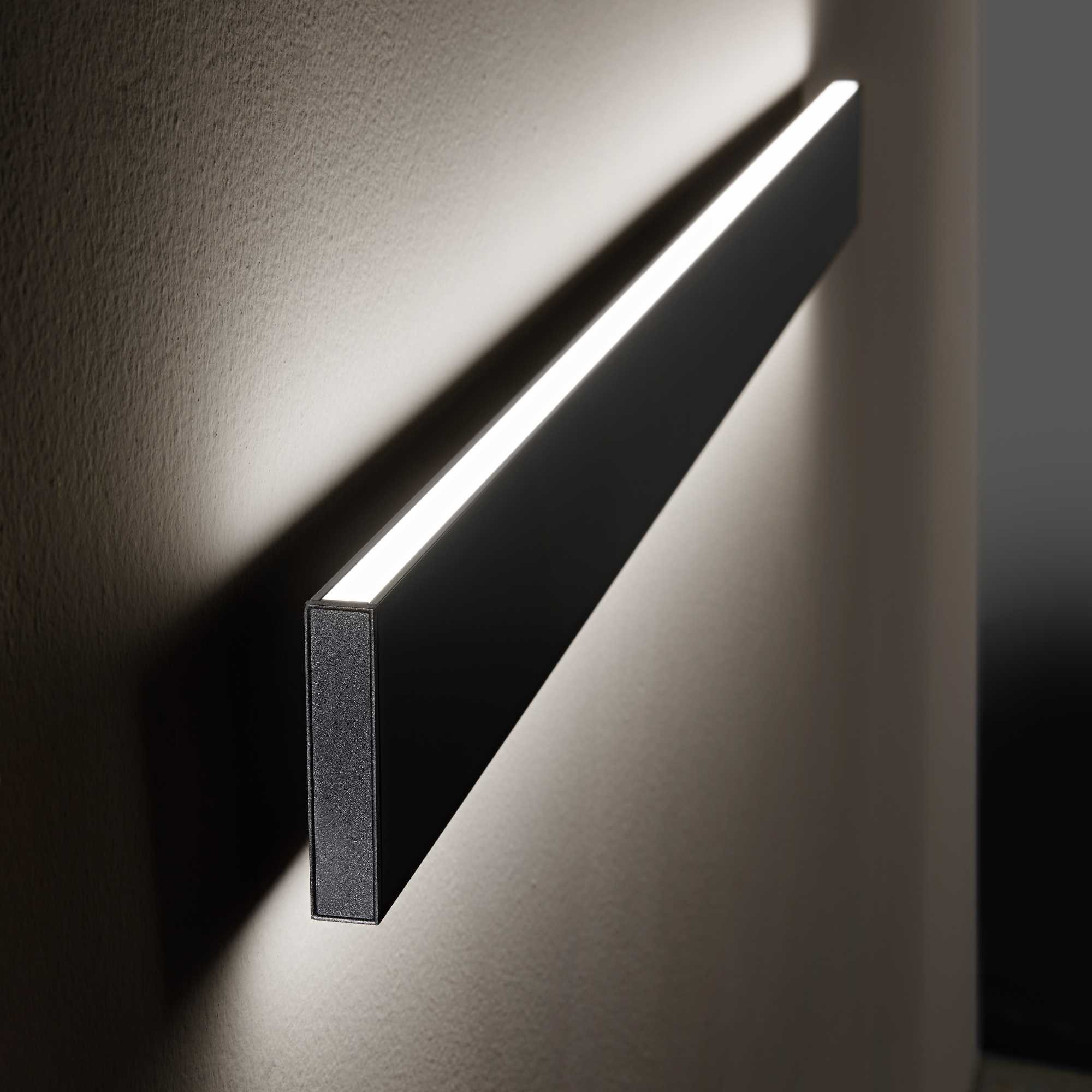 Una lampada da parete rettangolare nera con una luce bianca brillante. La lampada è montata su una parete chiara e strutturata.