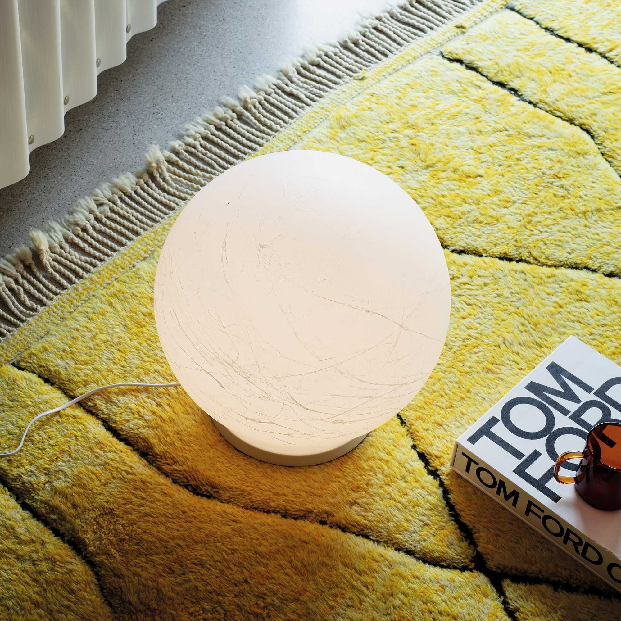 Una lampada sferica è su un tappeto giallo vicino a un libro e una tazza. Sullo sfondo c'è un muro bianco.