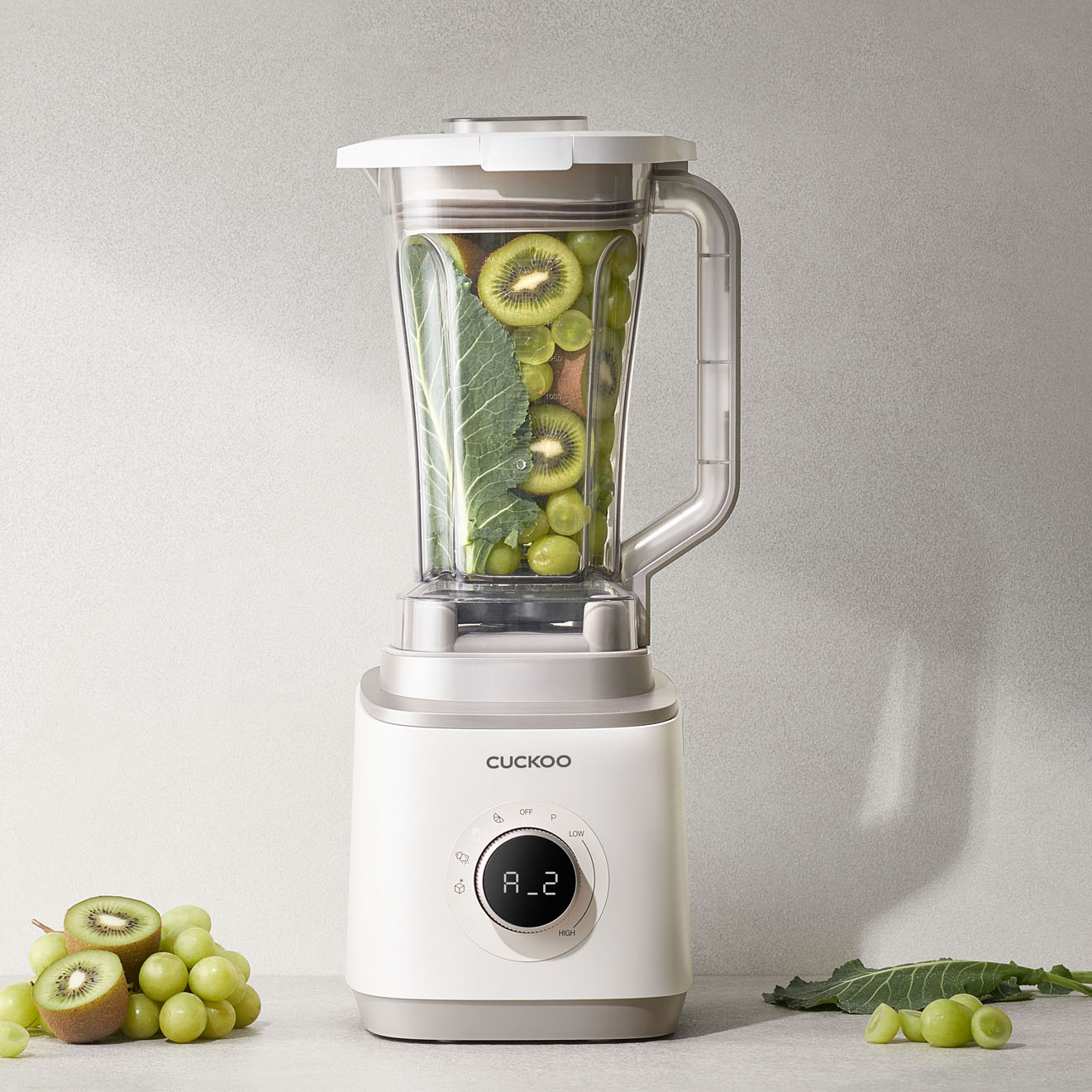 Ein weißer Mixer mit Kiwi und Trauben darin. Trauben und Kiwi liegen daneben. Das Bedienfeld des Mixers zeigt 'A_2'.