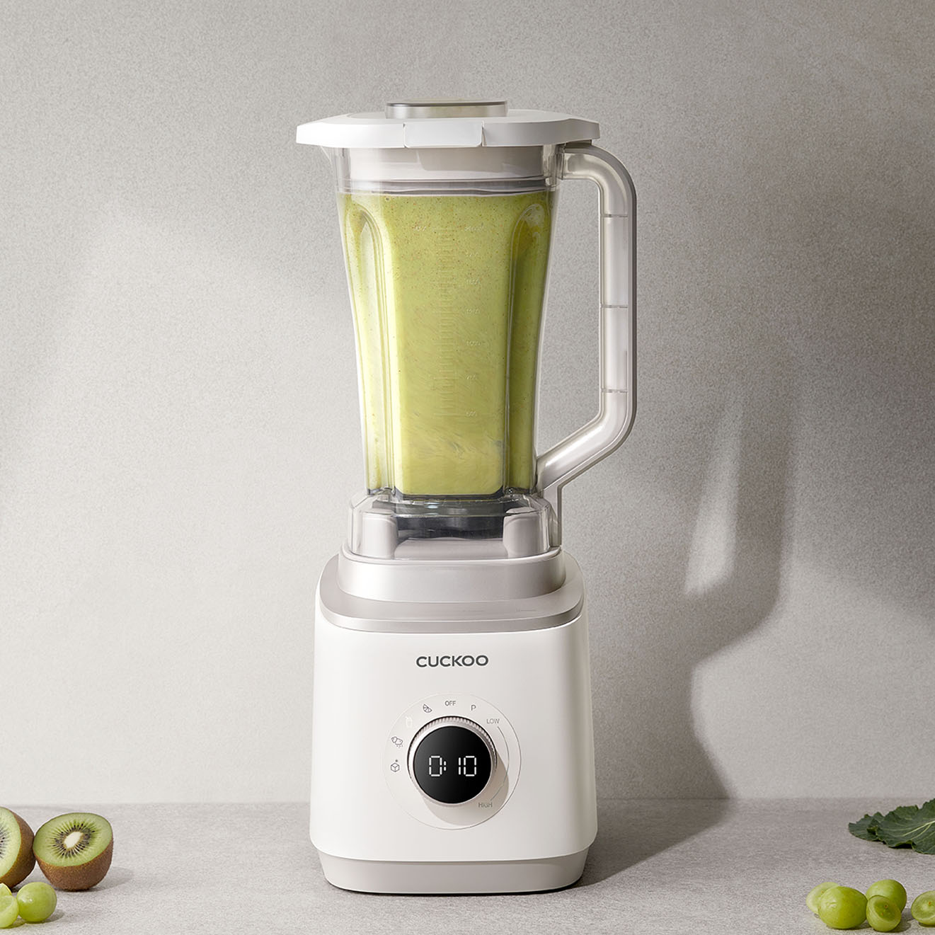 Ein weißer Mixer mit grünem Smoothie. Kiwi, Trauben und Grünkohl liegen daneben. Der Mixer zeigt '0:10' an.