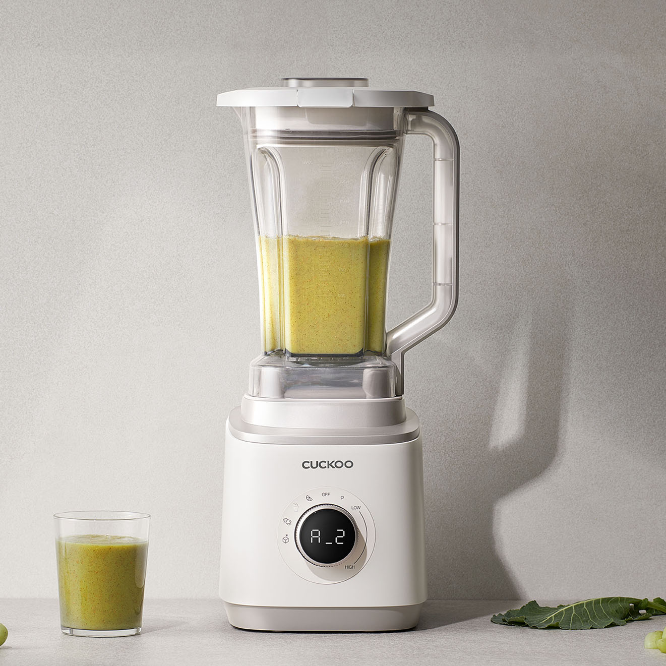 Ein weißer Cuckoo-Mixer mit grünem Smoothie. Ein Glas Smoothie steht daneben. Der Hintergrund ist eine graue Oberfläche.