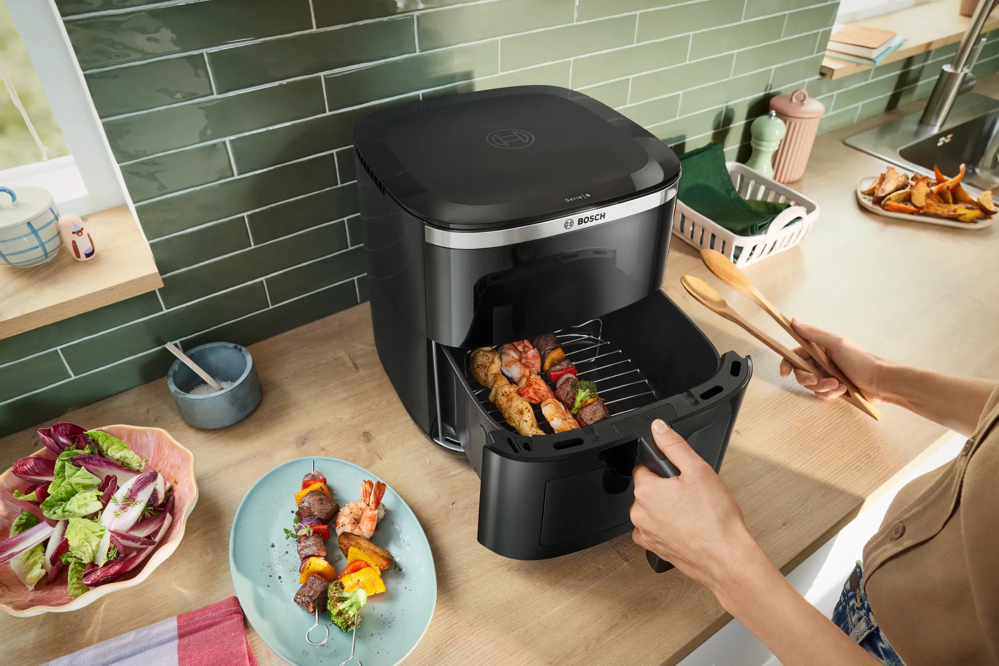 Frytownica powietrzna z szaszłykami w środku. Sałatka i szaszłyki na talerzach w pobliżu. Osoba trzyma drewniane łyżki.