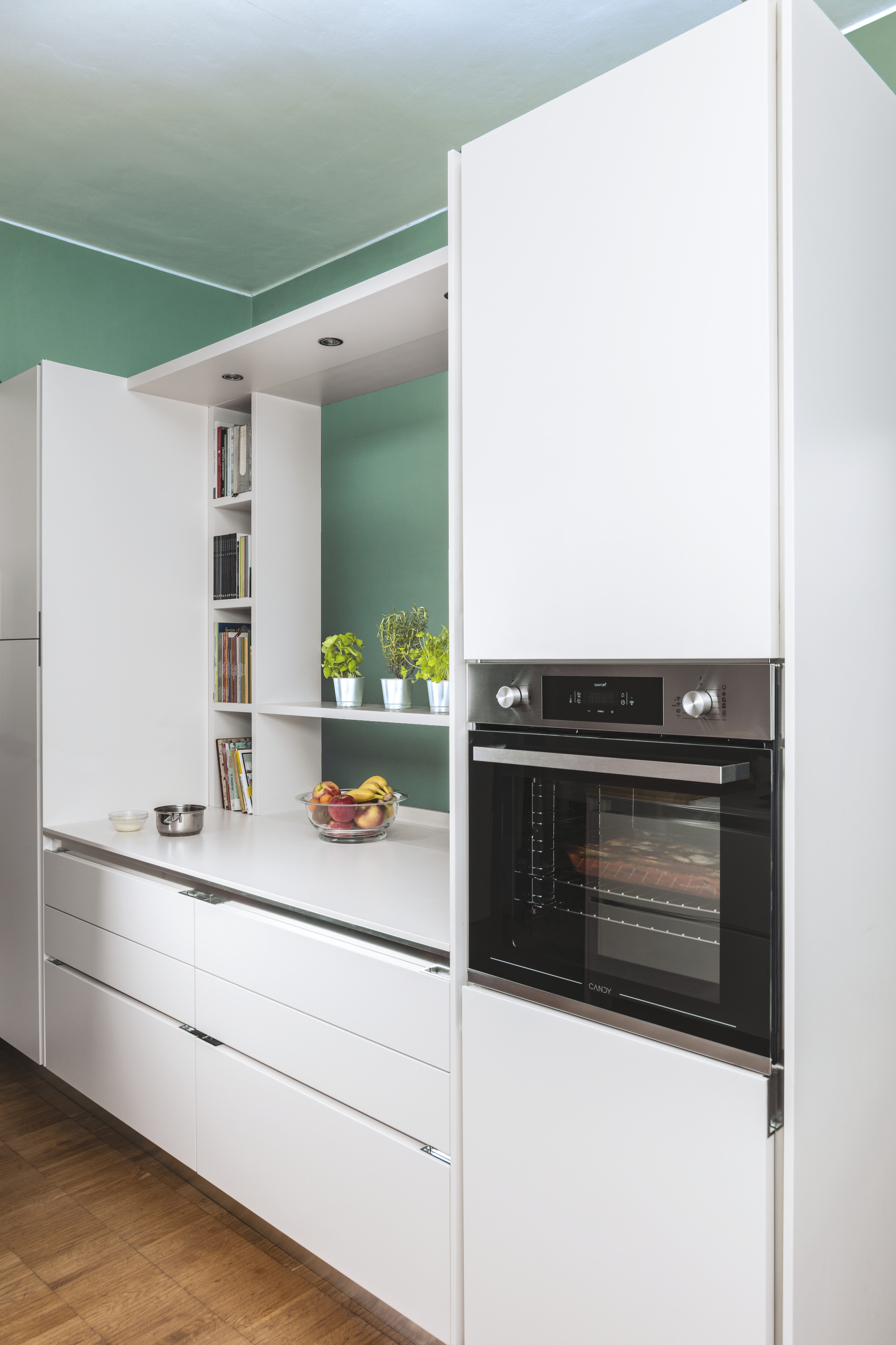 Cucina moderna con mobili bianchi, forno, fruttiera e piante. Parete verde sullo sfondo. Pavimento in legno.