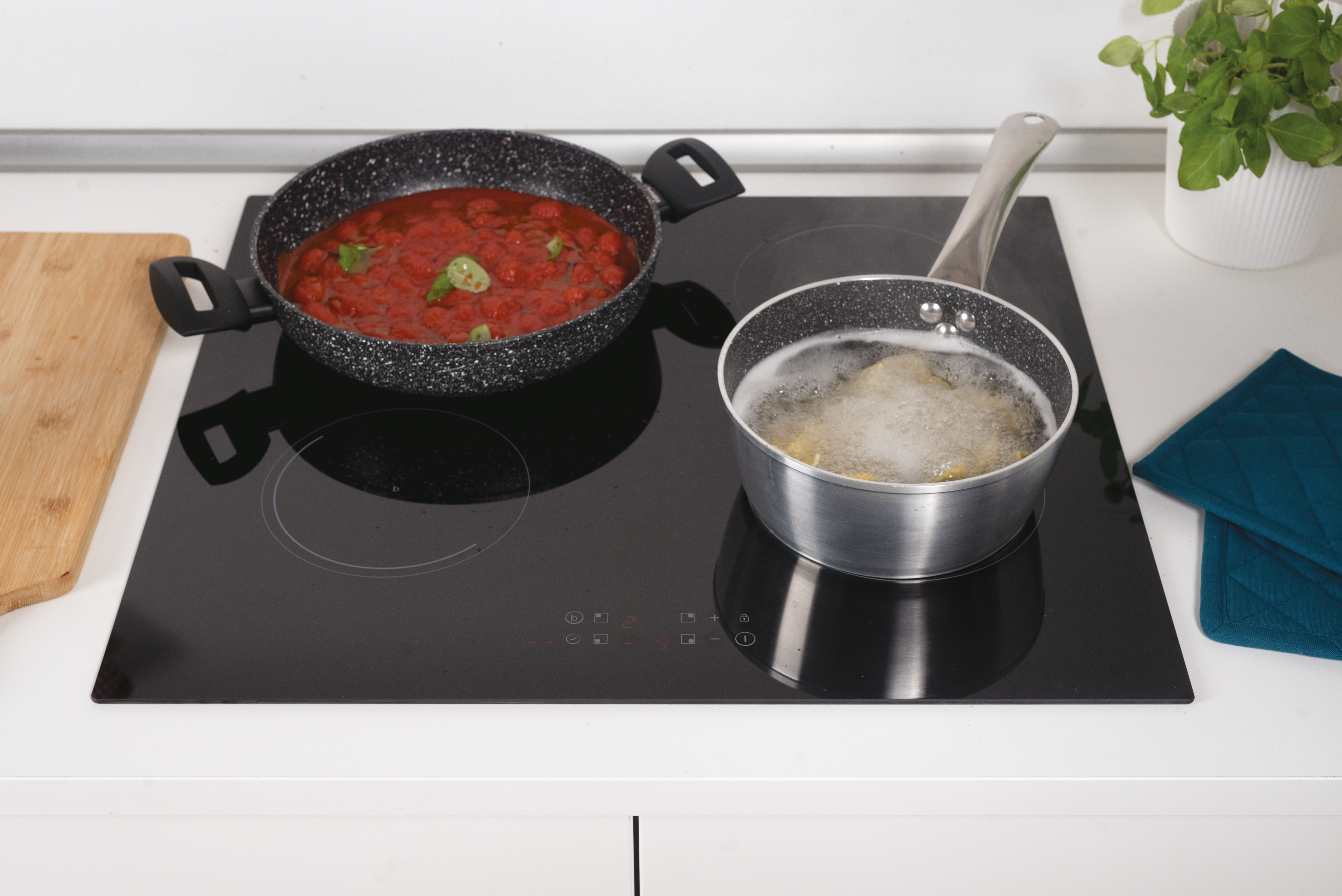 Czarna płyta kuchenna z dwoma garnkami: jednym z czerwonym sosem, drugim gotującym się. W pobliżu drewniana deska i turkusowe podkładki.