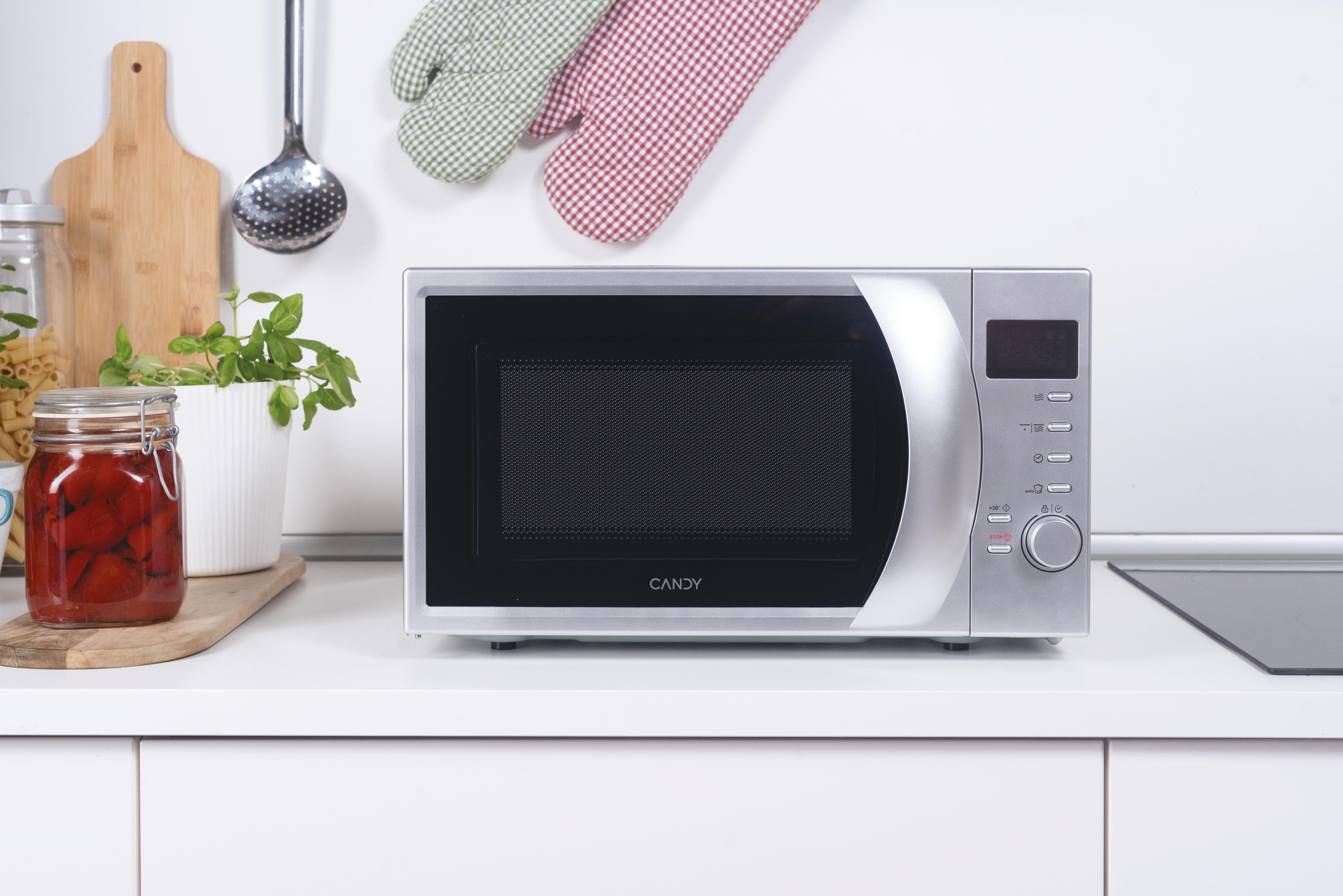 Un forno a microonde argentato su un bancone bianco. Utensili e ingredienti da cucina a sinistra. Guanti da forno appesi sopra.