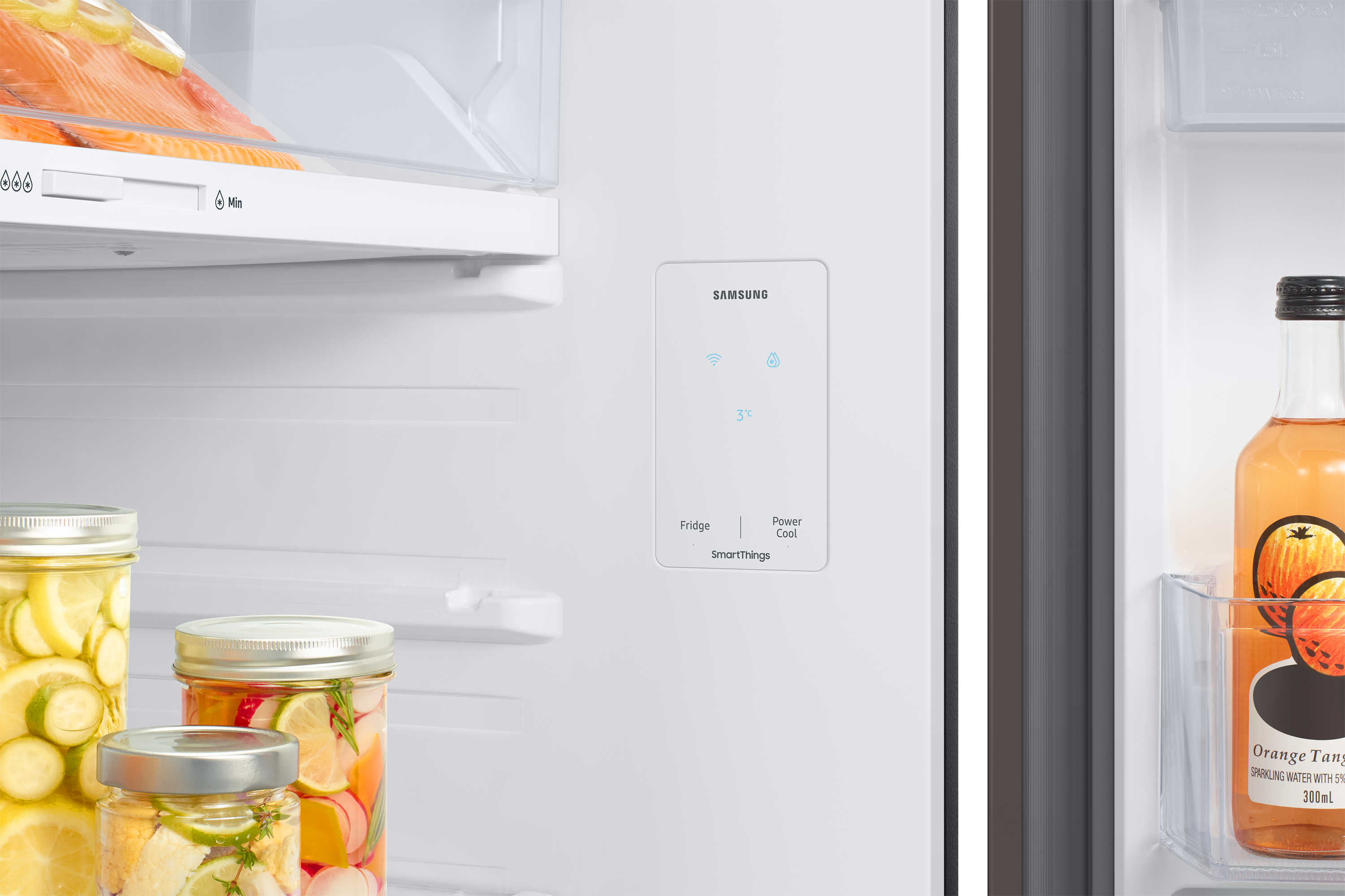 All'interno di un frigorifero sono visibili barattoli di cibo. Un pannello di controllo Samsung mostra 3°C. Una bottiglia di bevanda all'arancia è nella porta.