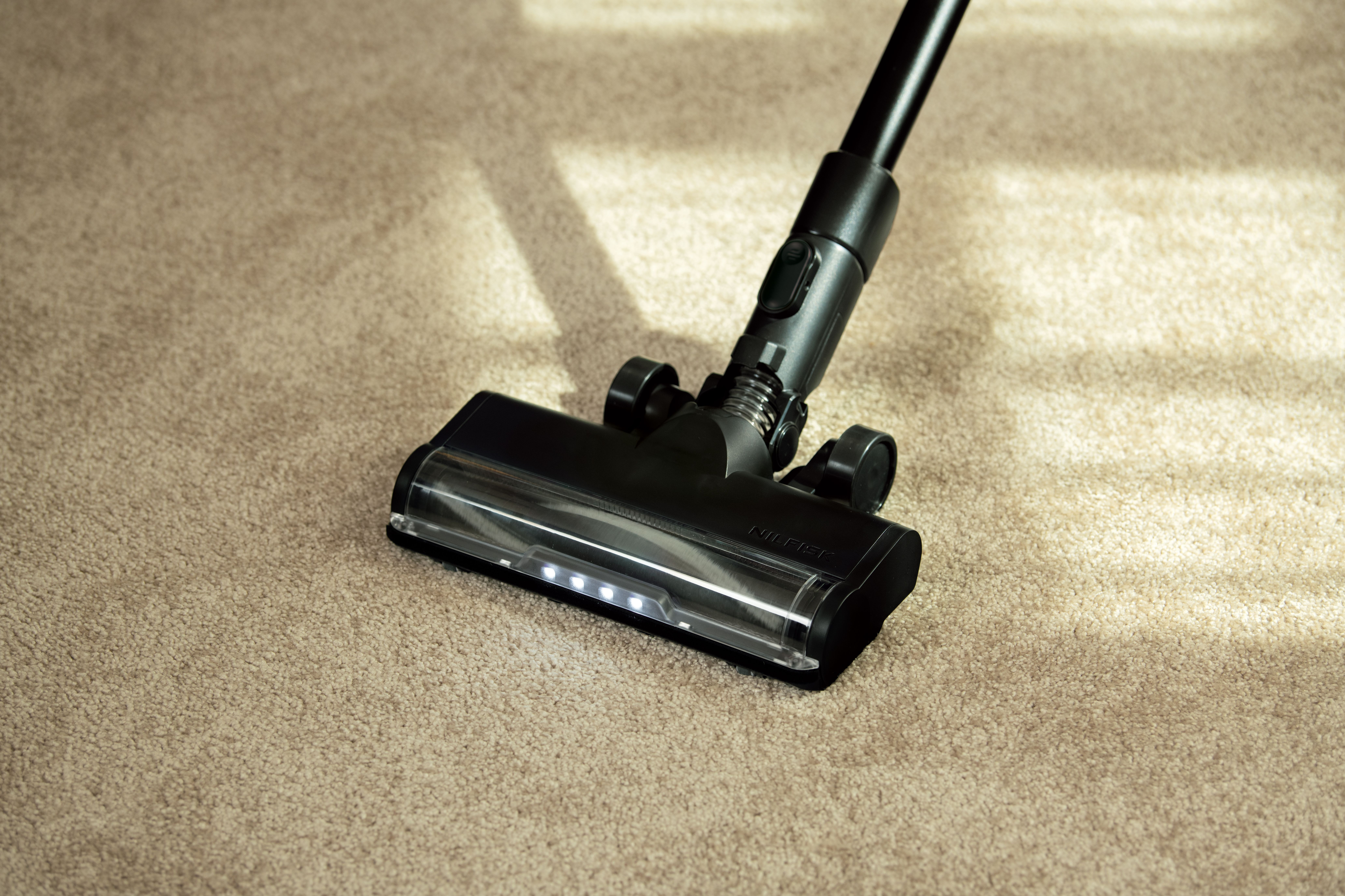 Une tête d'aspirateur noire sur un tapis beige. L'aspirateur est incliné, montrant sa brosse et quelques lumières.