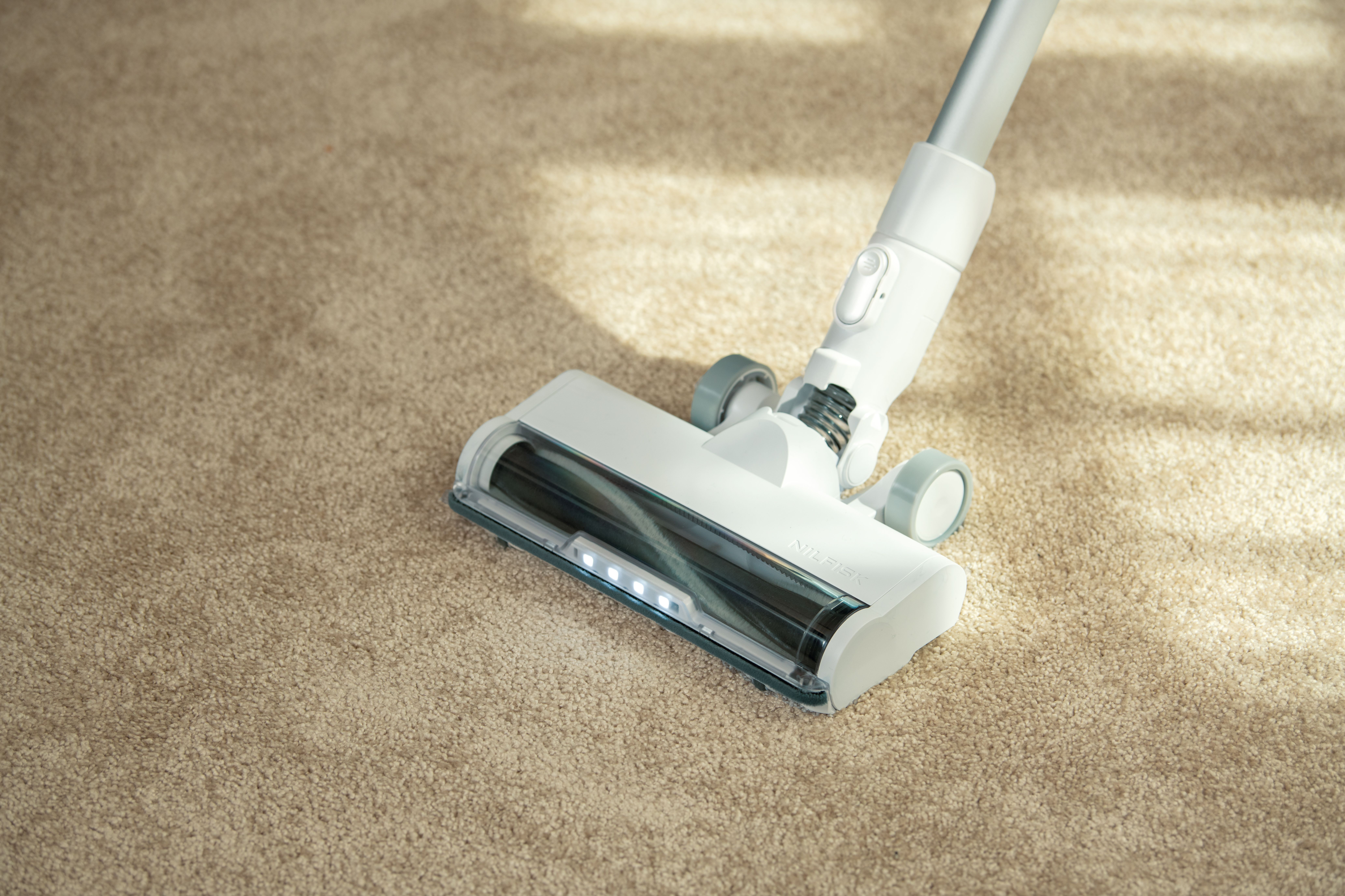 Una aspiradora blanca sobre una alfombra beige. El cabezal de la aspiradora está cerca del espectador, con el mango extendido hacia arriba.