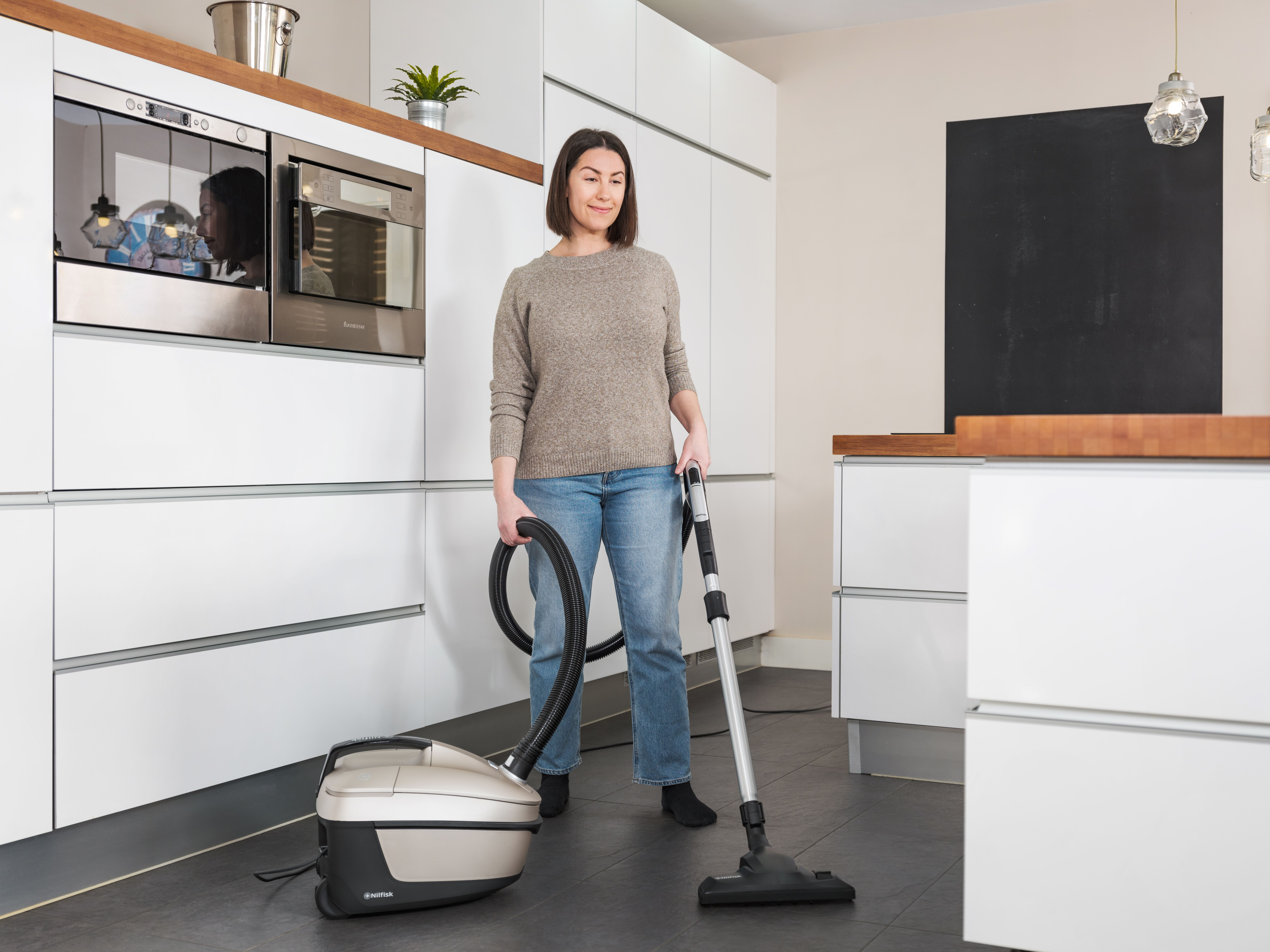 A woman vacuums a kitchen. She wears a sweater and jeans. A vacuum cleaner is on the floor. White cabinets are on the walls.