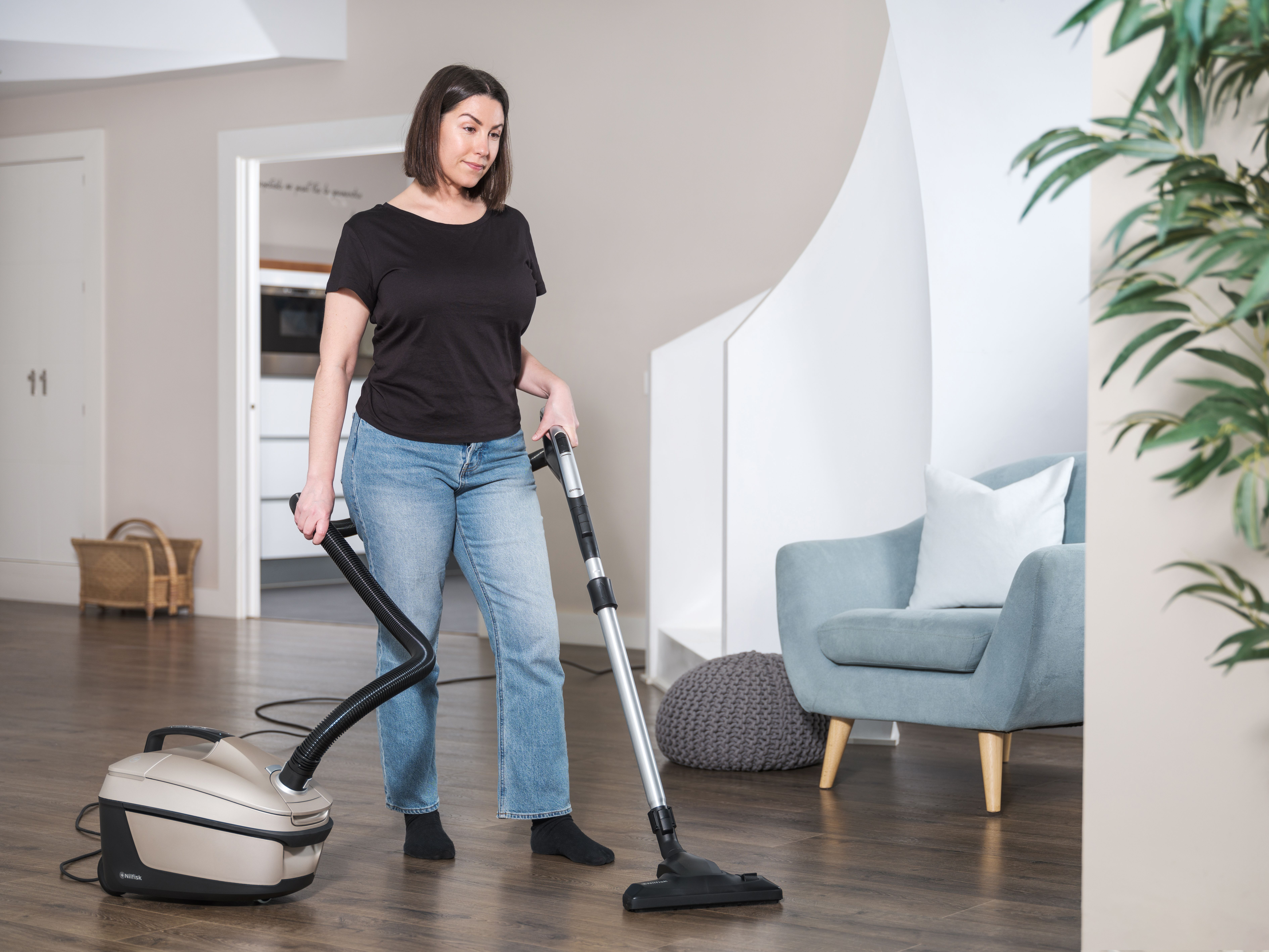 A woman vacuums a wooden floor. She wears jeans and a black shirt. Furniture and plants are in the background.