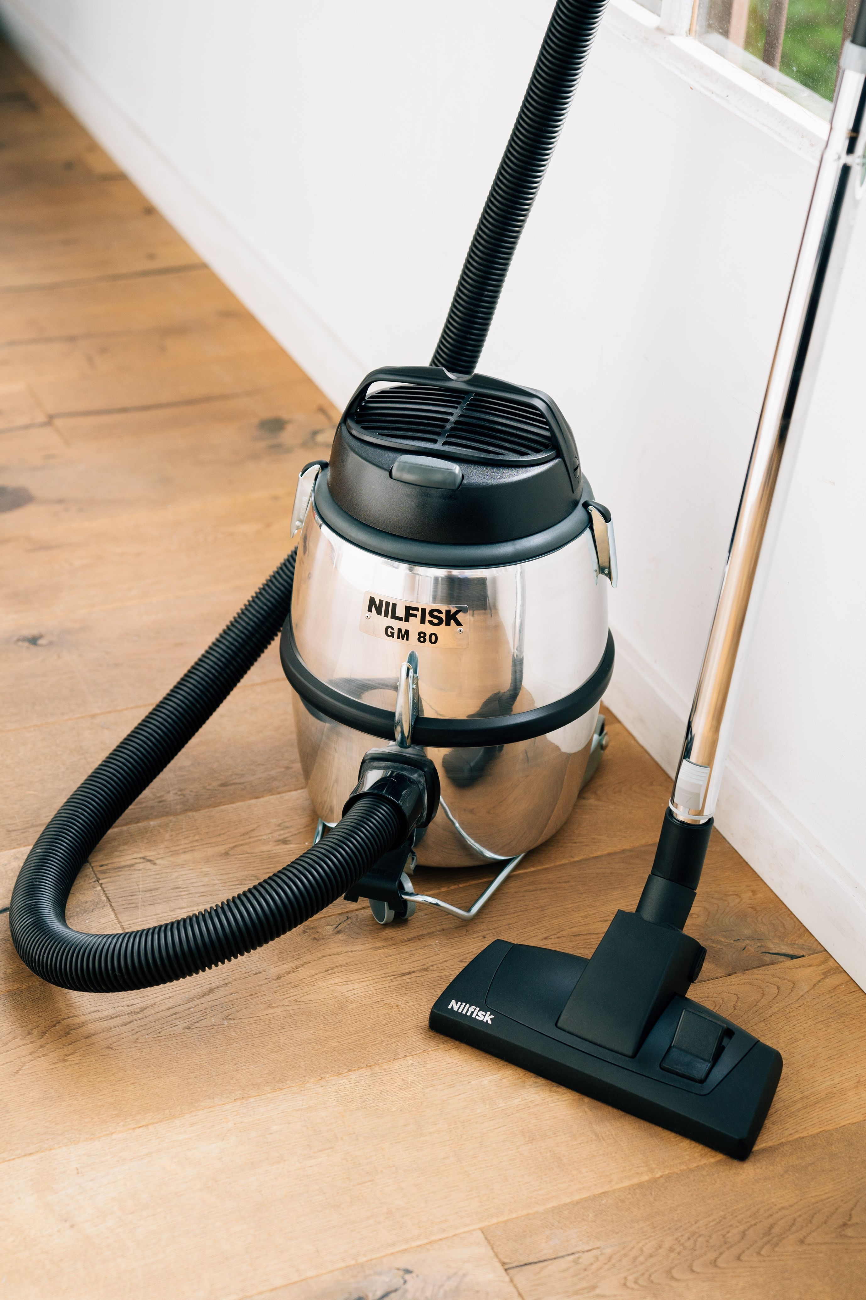 Un aspirateur argenté Nilfisk GM 80 avec tuyau et tête noirs est sur un plancher en bois.
