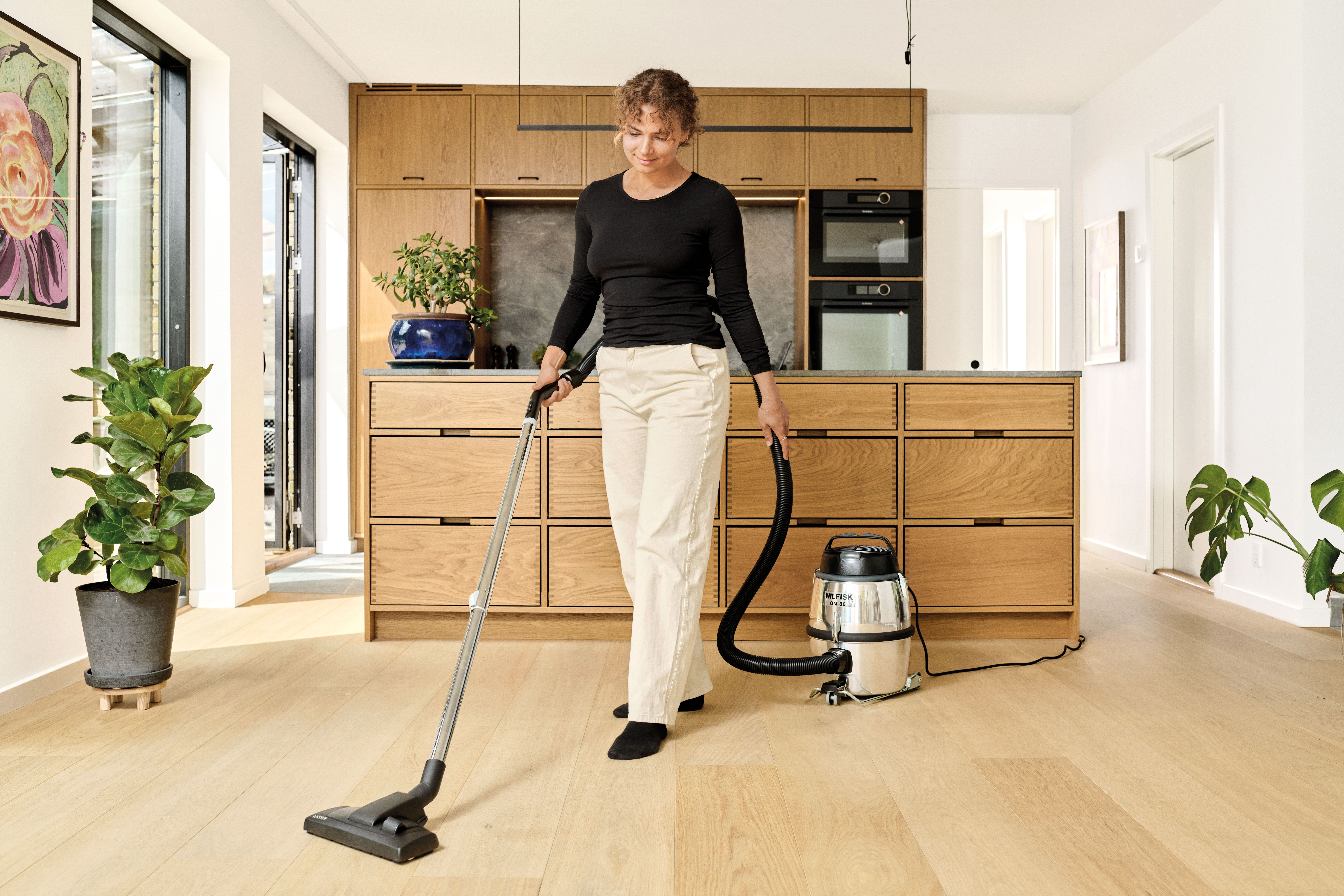 Eine Frau saugt in einer modernen Küche mit Holzschränken und einem silbernen Staubsauger einen hellen Holzboden.