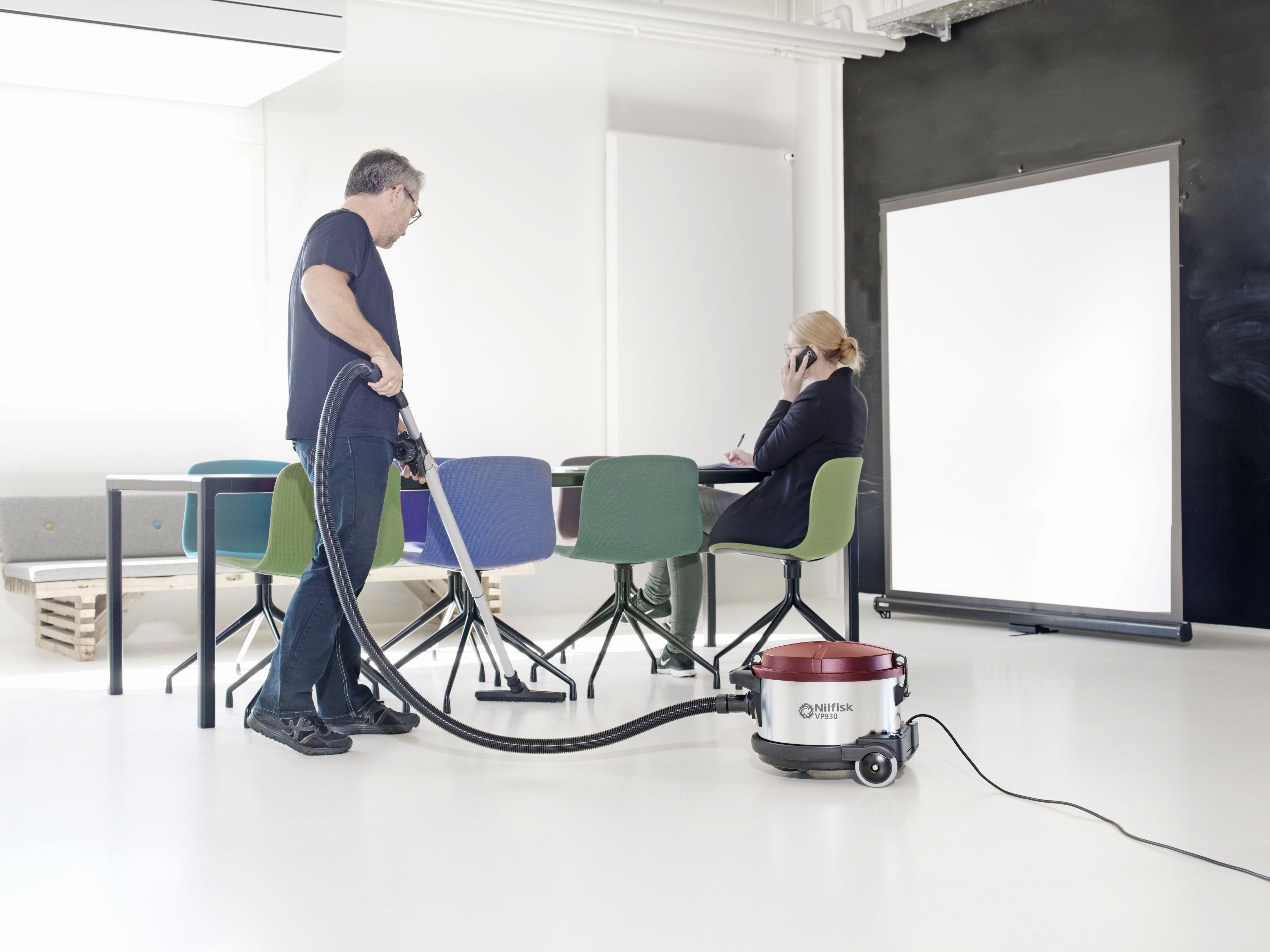 Mężczyzna odkurza biuro. Kobieta rozmawia przez telefon przy stole. Odkurzacz jest czerwono-srebrny. Krzesła są w różnych kolorach.