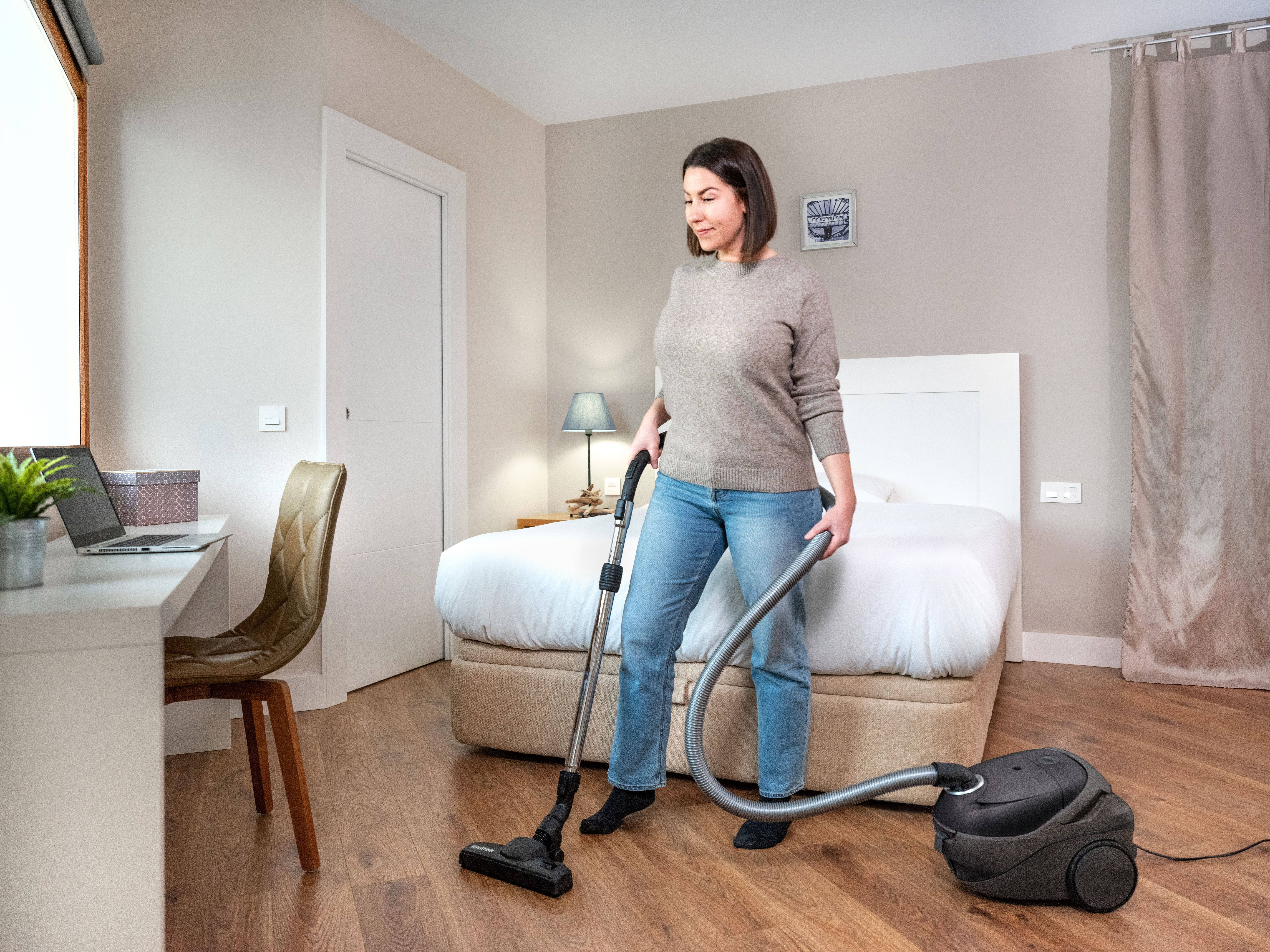 Una mujer aspira una habitación. Hay una cama detrás. A la izquierda hay un escritorio con un portátil y una silla.
