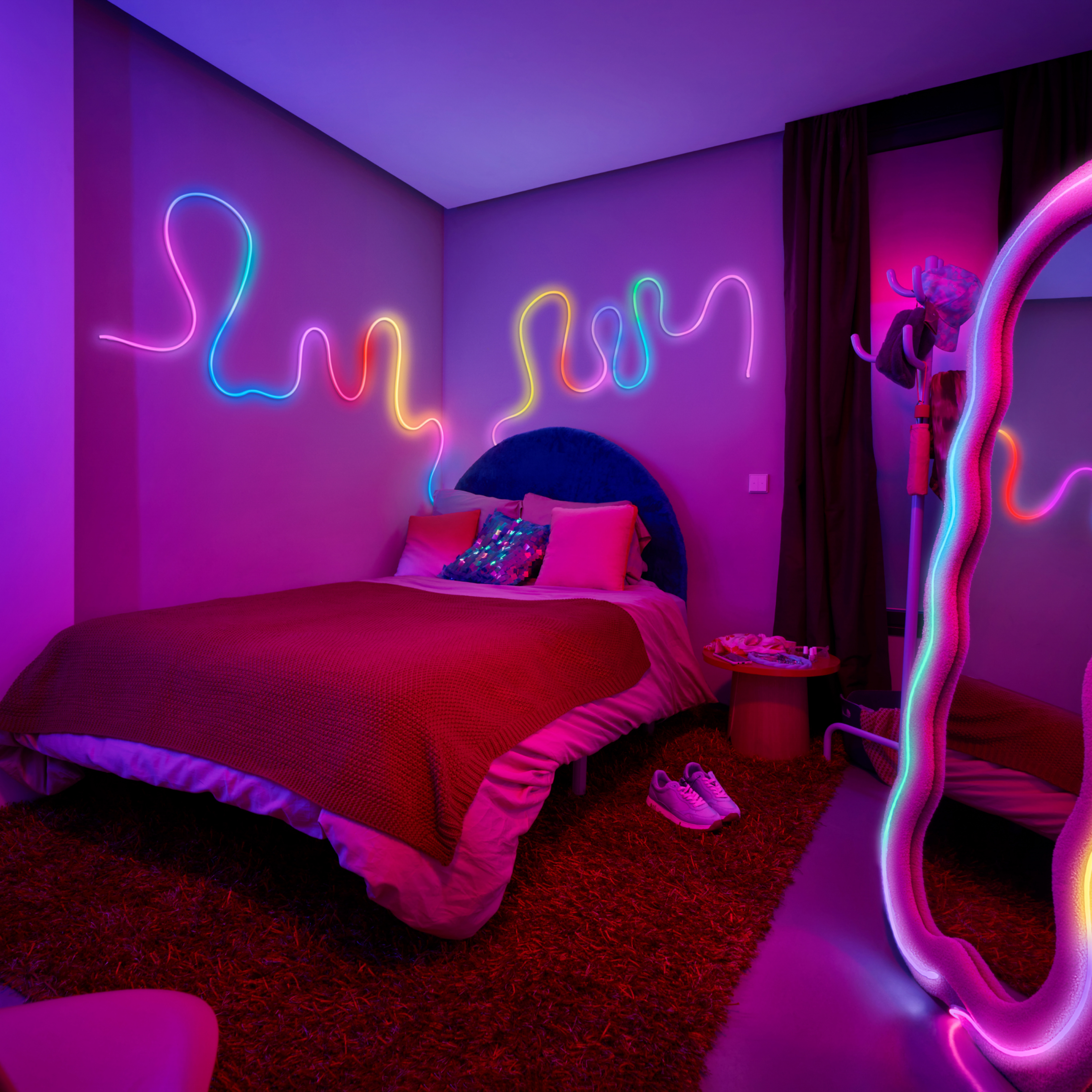 Bedroom with neon lights. Bed with red blanket, pink pillows, and shoes on the floor. Mirror with neon.