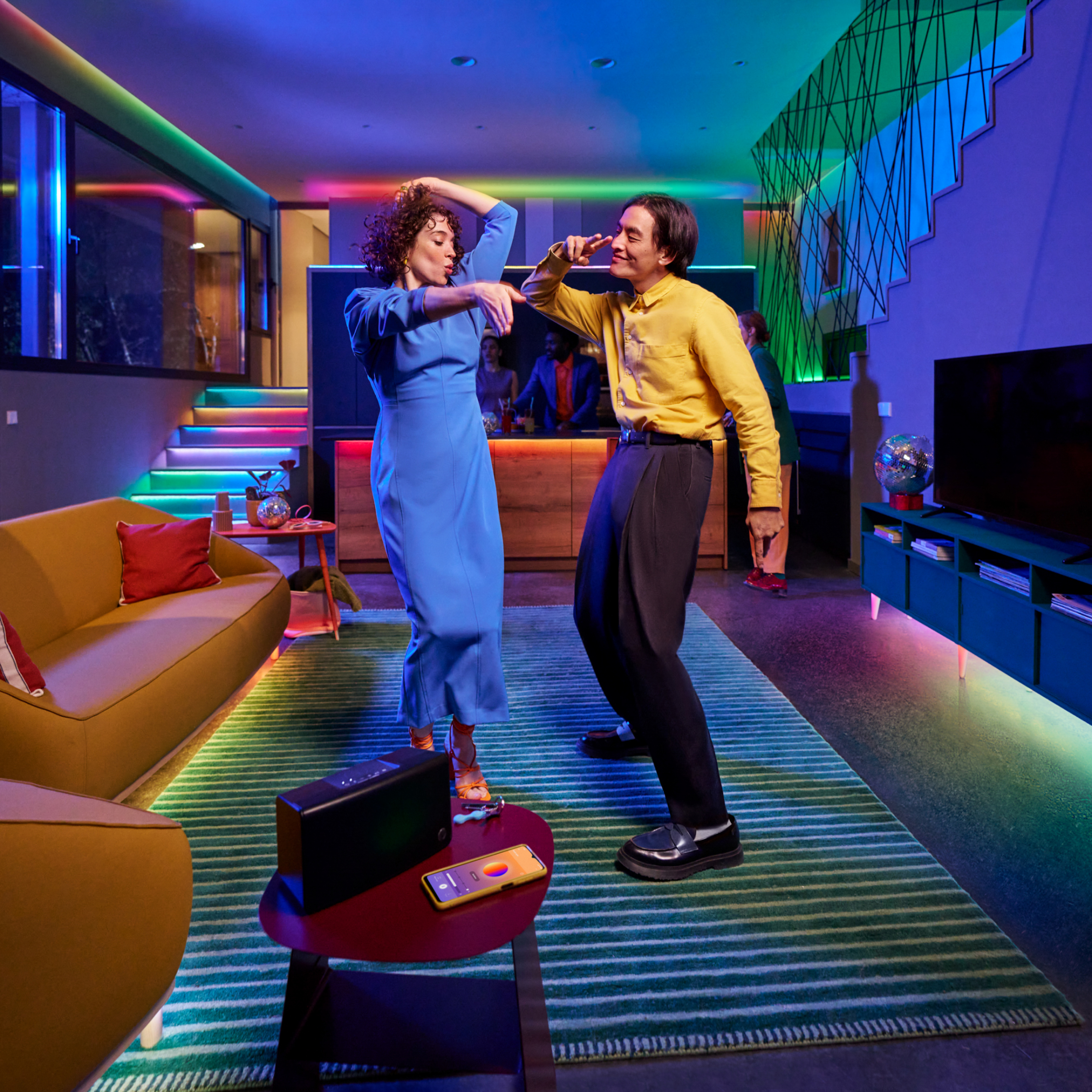 Two people dance in a room with colorful lights. A speaker and phone sit on a table. A bar and stairs are in the background.