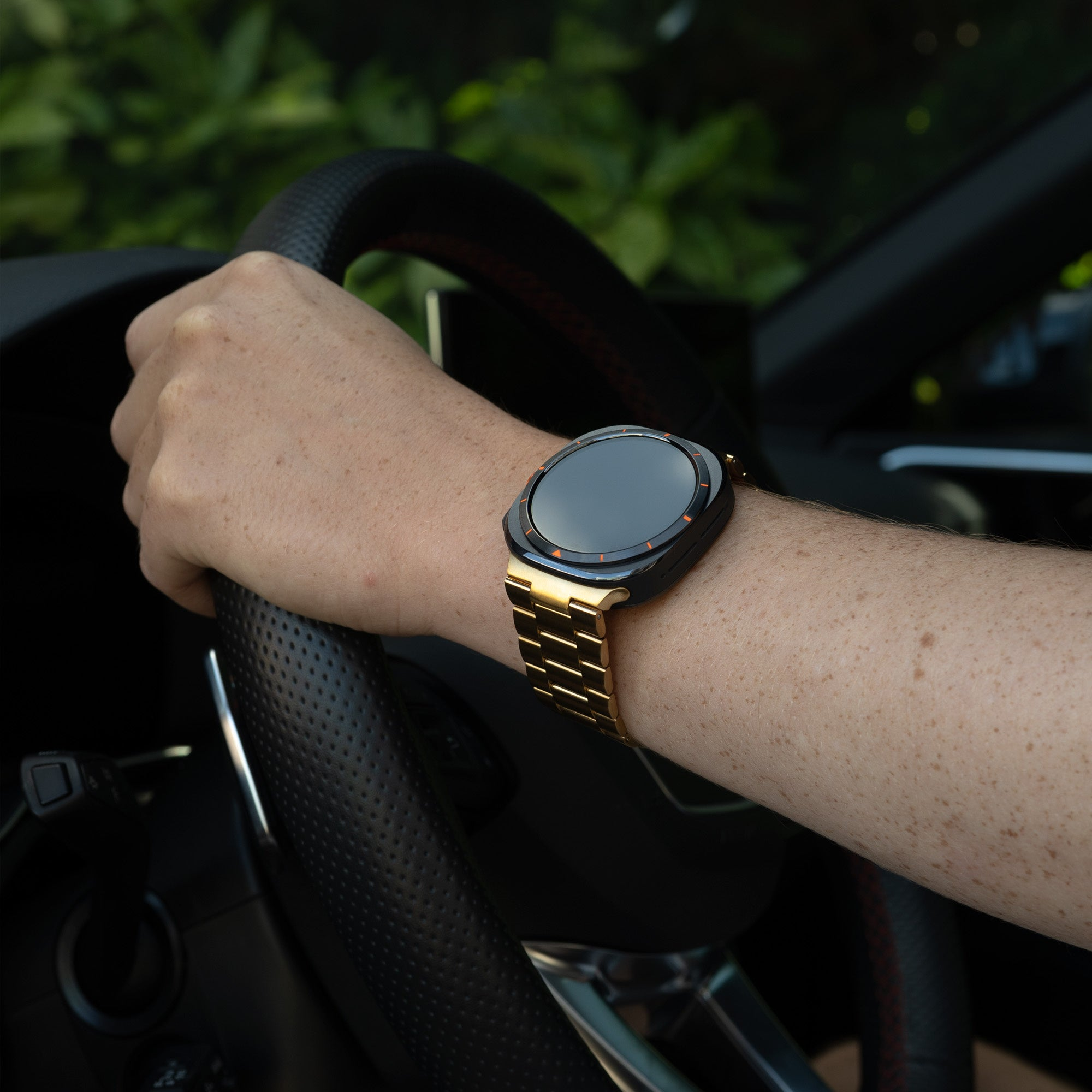 Ein Arm hält ein Lenkrad. Am Handgelenk befindet sich eine goldene Uhr. Der Hintergrund ist verschwommen.