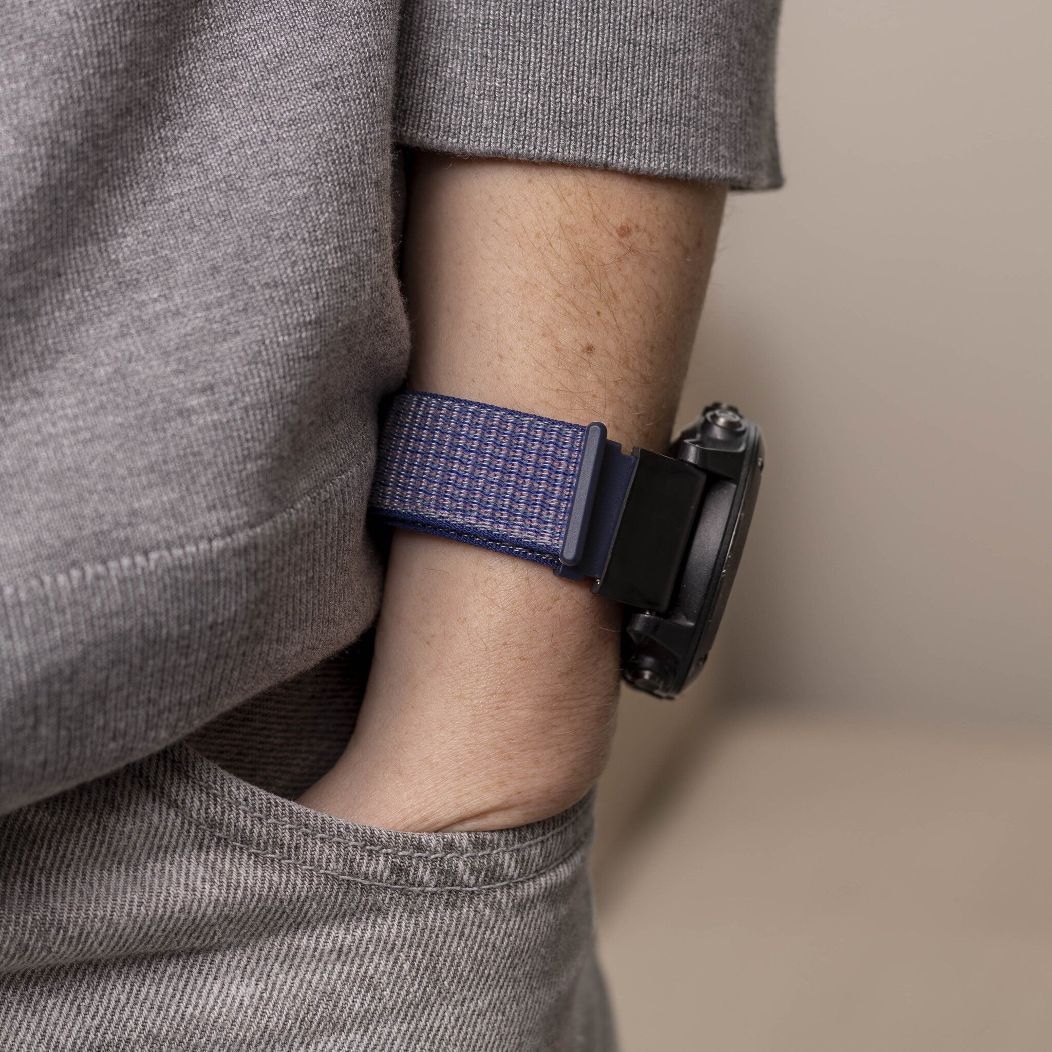 Person trägt einen grauen Pullover, eine Uhr mit blauem Armband. Arm in der Tasche. Beigefarbener Hintergrund.