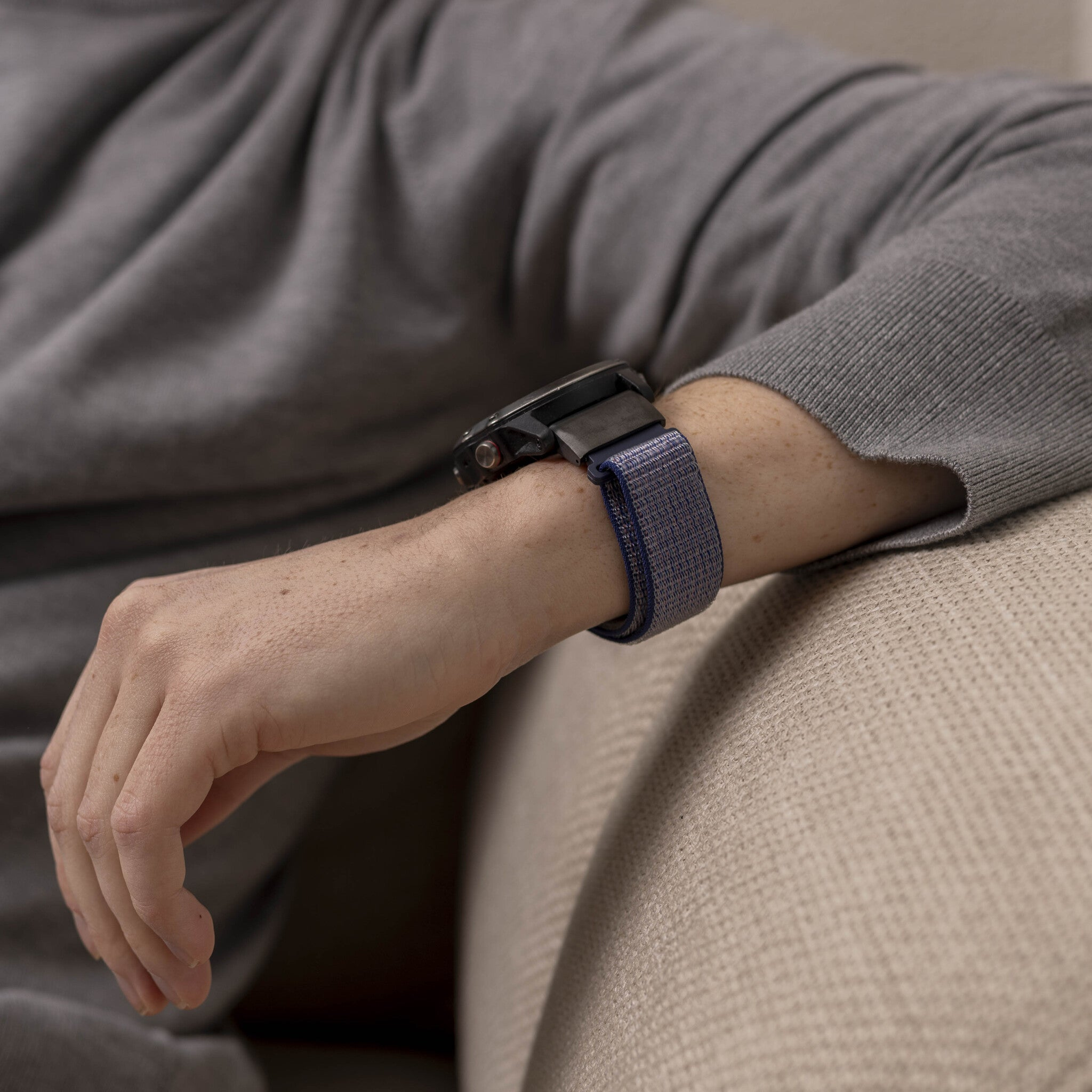 Handgelenk einer Person mit Uhr. Die Uhr hat ein blau-graues Stoffarmband. Die Person trägt einen grauen Pullover.