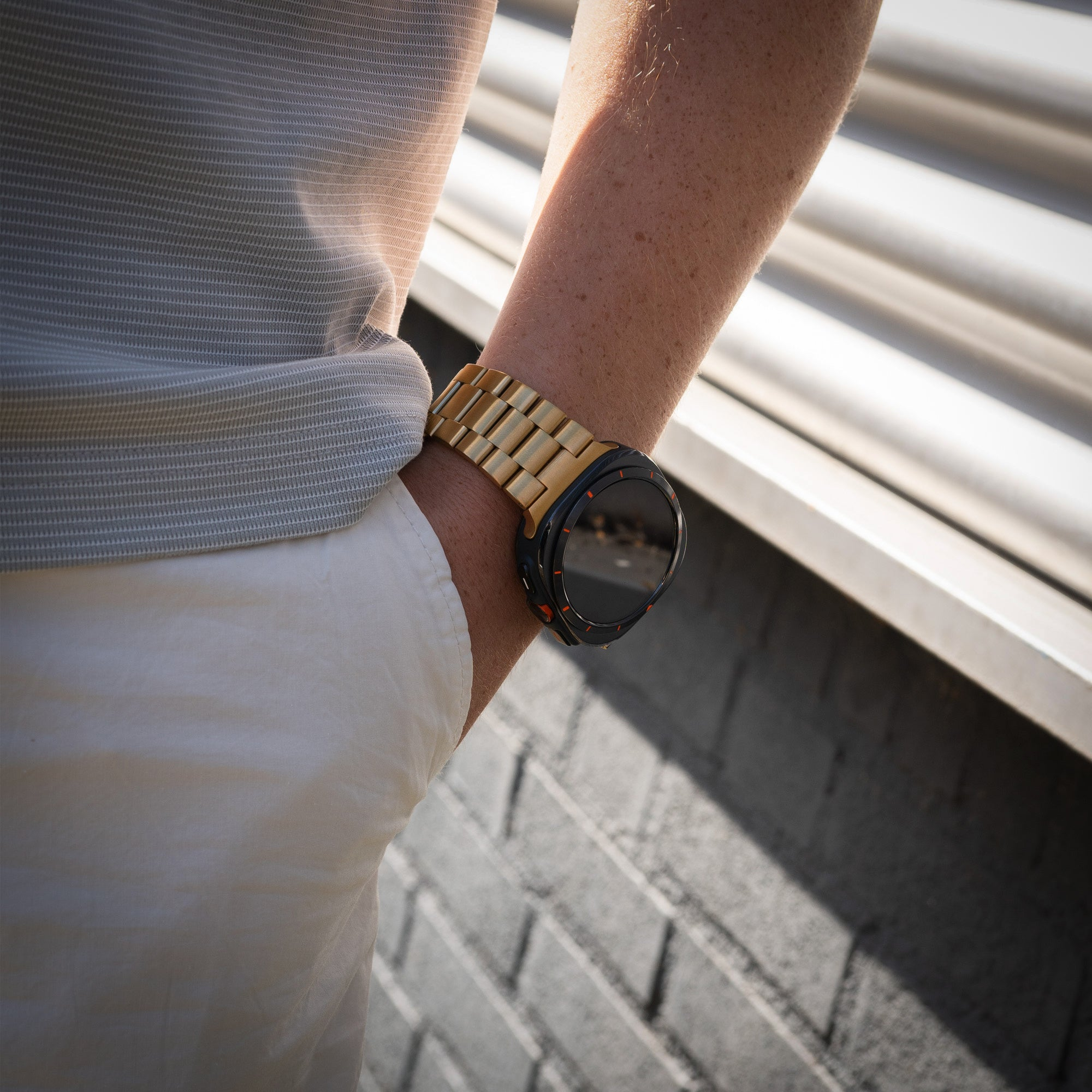 Ein Arm mit einer goldenen Uhr. Die Person trägt ein graues Hemd und eine weiße Hose. Der Hintergrund ist eine Backsteinmauer.