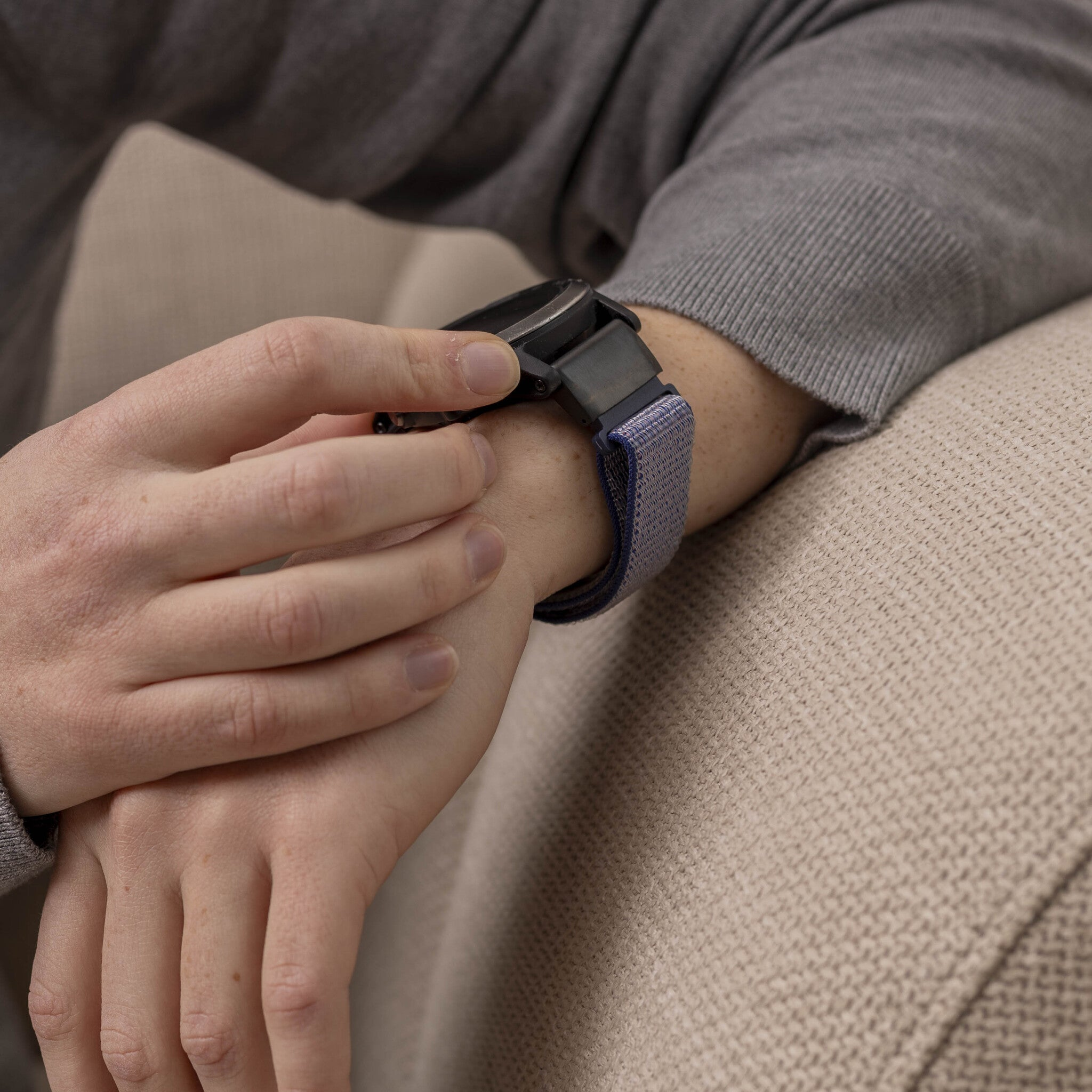 Eine Person in grauem Pullover, die eine schwarze Smartwatch mit blauem Armband überprüft.