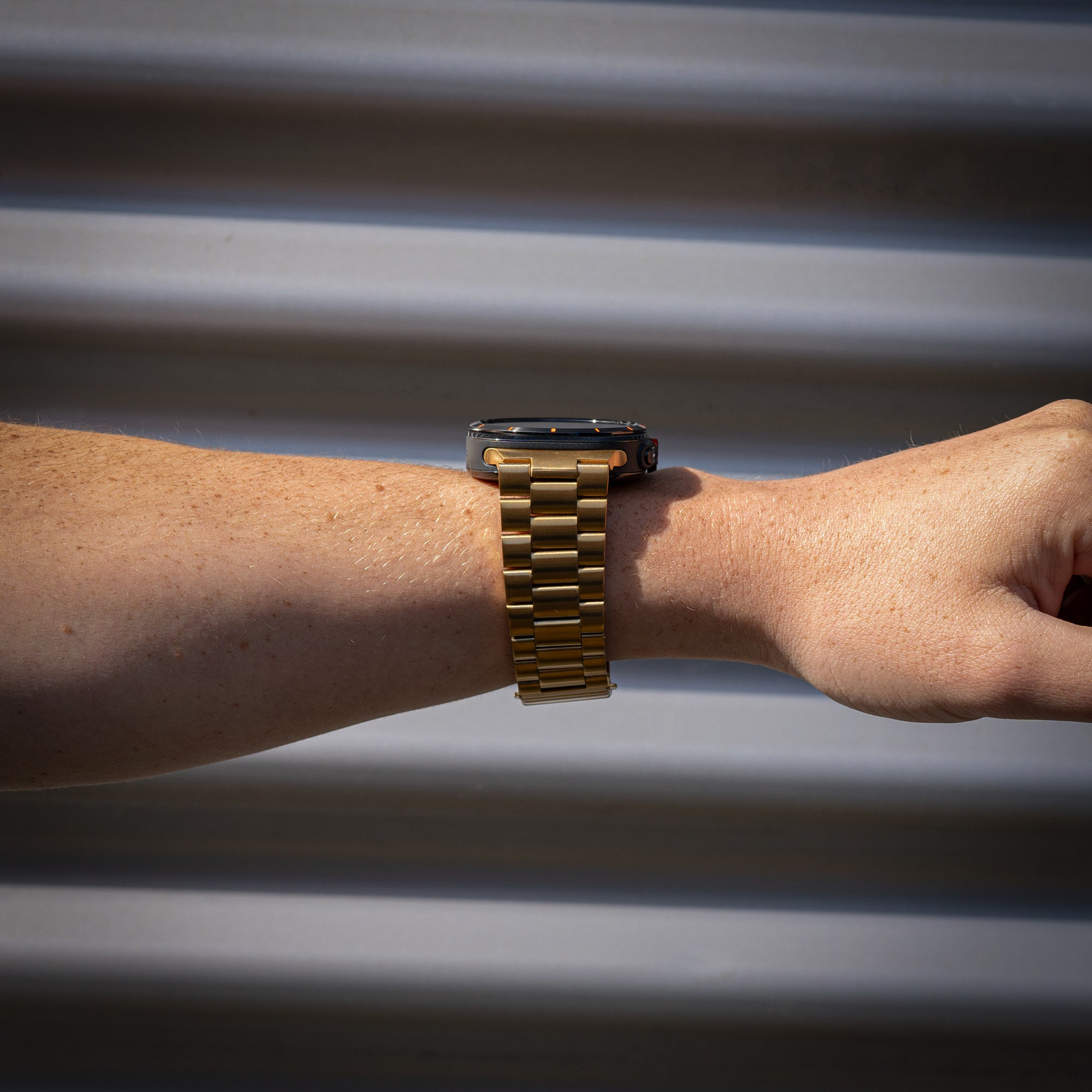 Ein Handgelenk mit einer goldenen Uhr. Die Haut hat Sommersprossen. Der Hintergrund ist eine Metalloberfläche.