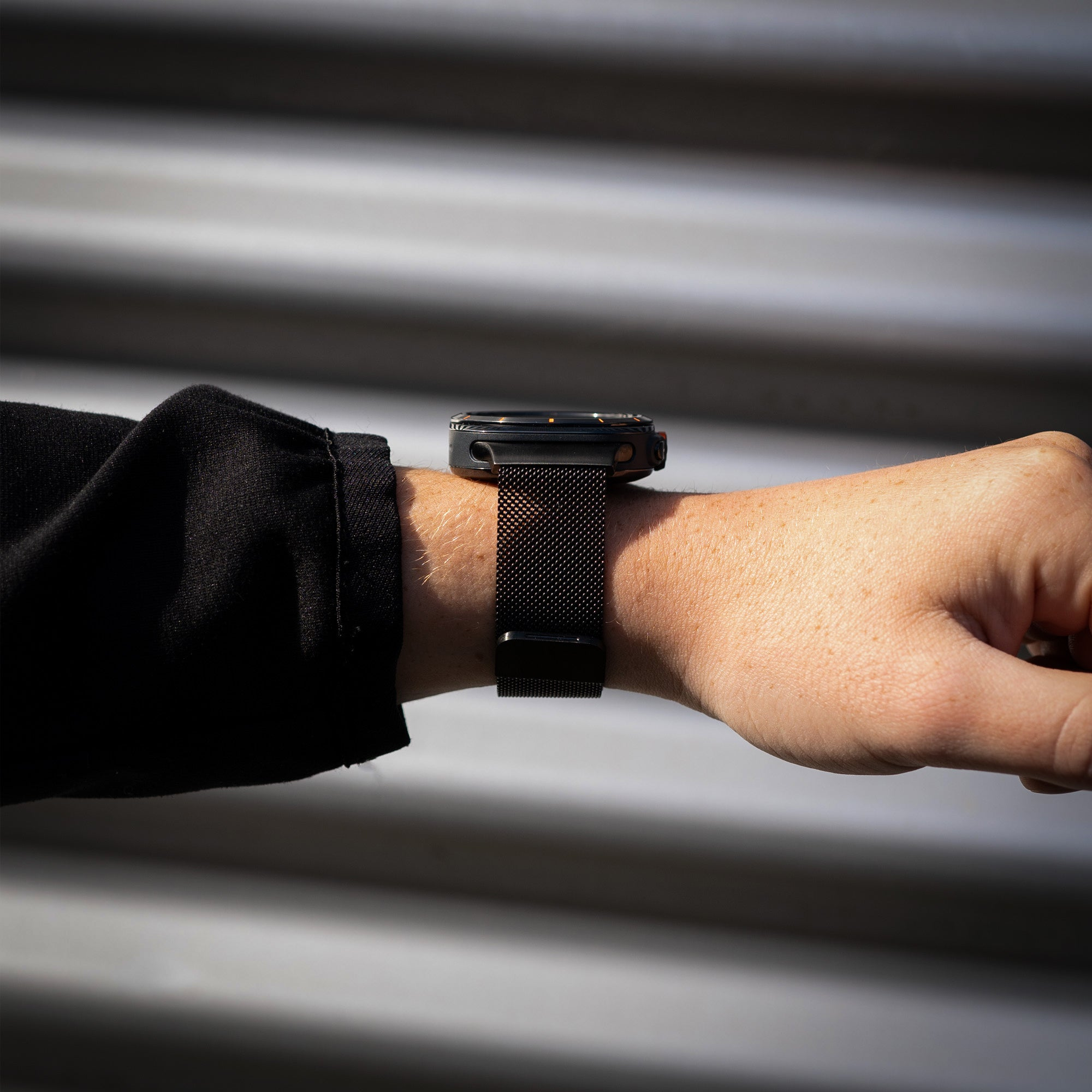 Ein Handgelenk mit einer schwarzen Smartwatch und einem schwarzen Metallarmband. Der Hintergrund ist eine metallische, gestreifte Wand.