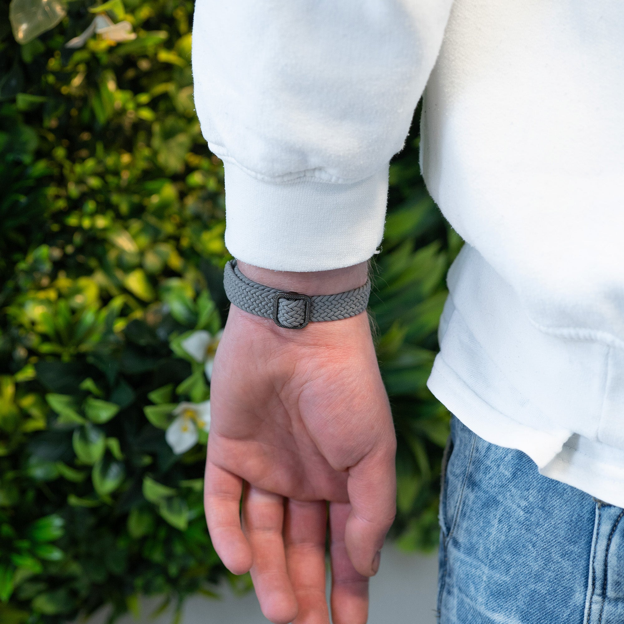 Eine Person, die ein graues geflochtenes Armband mit schwarzer Schnalle trägt. Die Person trägt ein weißes Hemd und Jeans.