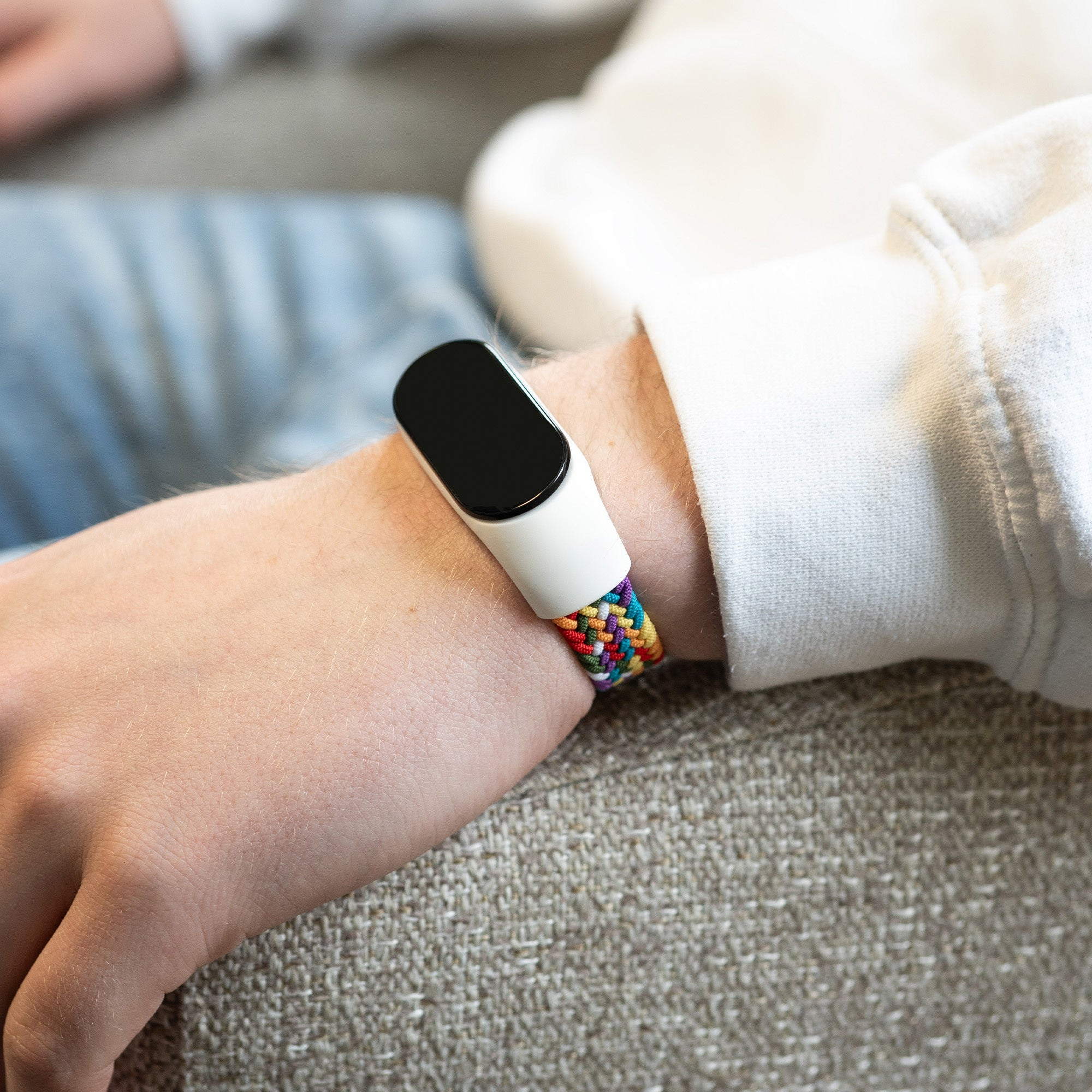 Handgelenk einer Person mit weißer Smartwatch und buntem geflochtenem Armband. Der Hintergrund ist unscharf.